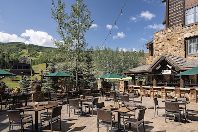 This image showcases an inviting outdoor patio area, perfect for dining or relaxation. The patio features multiple tables and chairs arranged neatly on a paved surface. A rustic bar area provides additional seating and ambiance, set against a backdrop of trees and mountains.