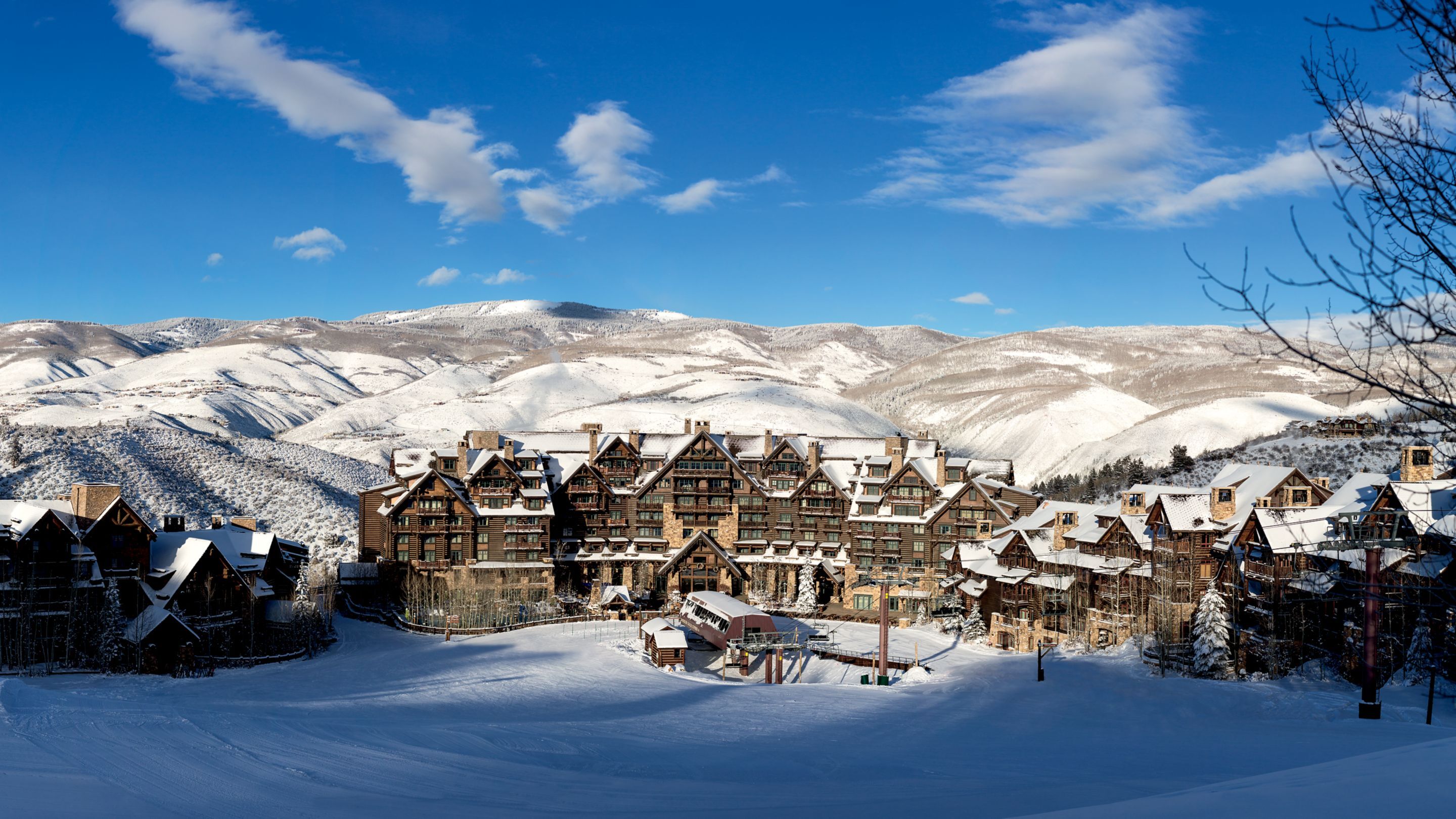 This is an exterior front view of a large, luxurious ski resort or lodge nestled in a snowy mountain landscape. The architecture features a rustic, multi-story structure with wood and stone elements and multiple gabled roofs. Snow covers all exterior surfaces, and a ski lift is visible, enhancing the winter resort atmosphere.