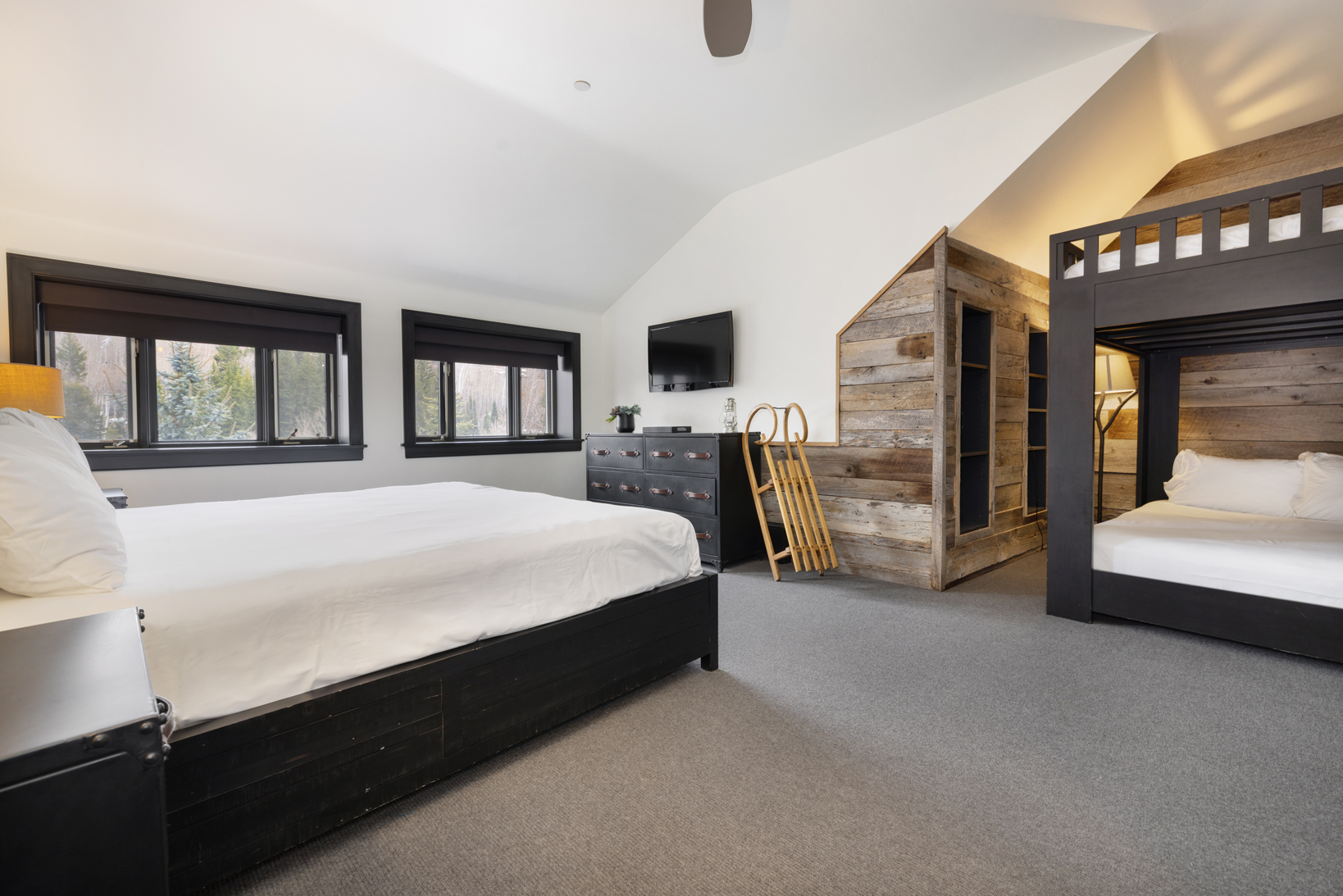 The image showcases a guest bedroom featuring a bed with a dark wood frame and white linens. A rustic wooden bunk bed set is also present, adding a unique touch to the room. The room benefits from natural light through the windows and also has a wooden closet feature built into the wall.