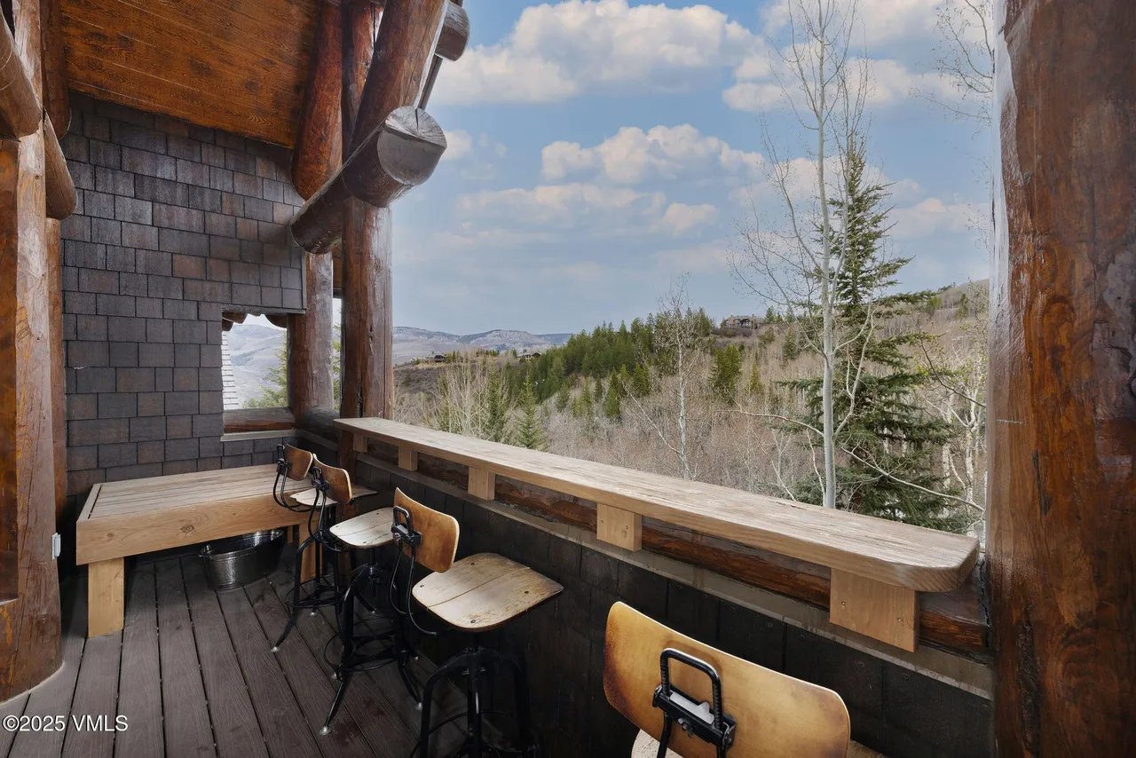 The image showcases a rustic balcony or deck area with wooden flooring and various seating options, including bar stools and a bench. The space is constructed with large wooden beams and features a view of a landscape with trees and mountains under a partly cloudy sky. The natural materials and open design suggest a relaxing outdoor living space.
