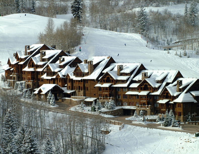 This image showcases the front exterior of a large, luxurious mountain lodge or condominium complex. The structure is characterized by its extensive use of wood, intricate rooflines covered in snow, and multiple levels. The setting is a snowy, mountainous landscape, suggesting a ski resort or winter retreat, which adds to the property's appeal.