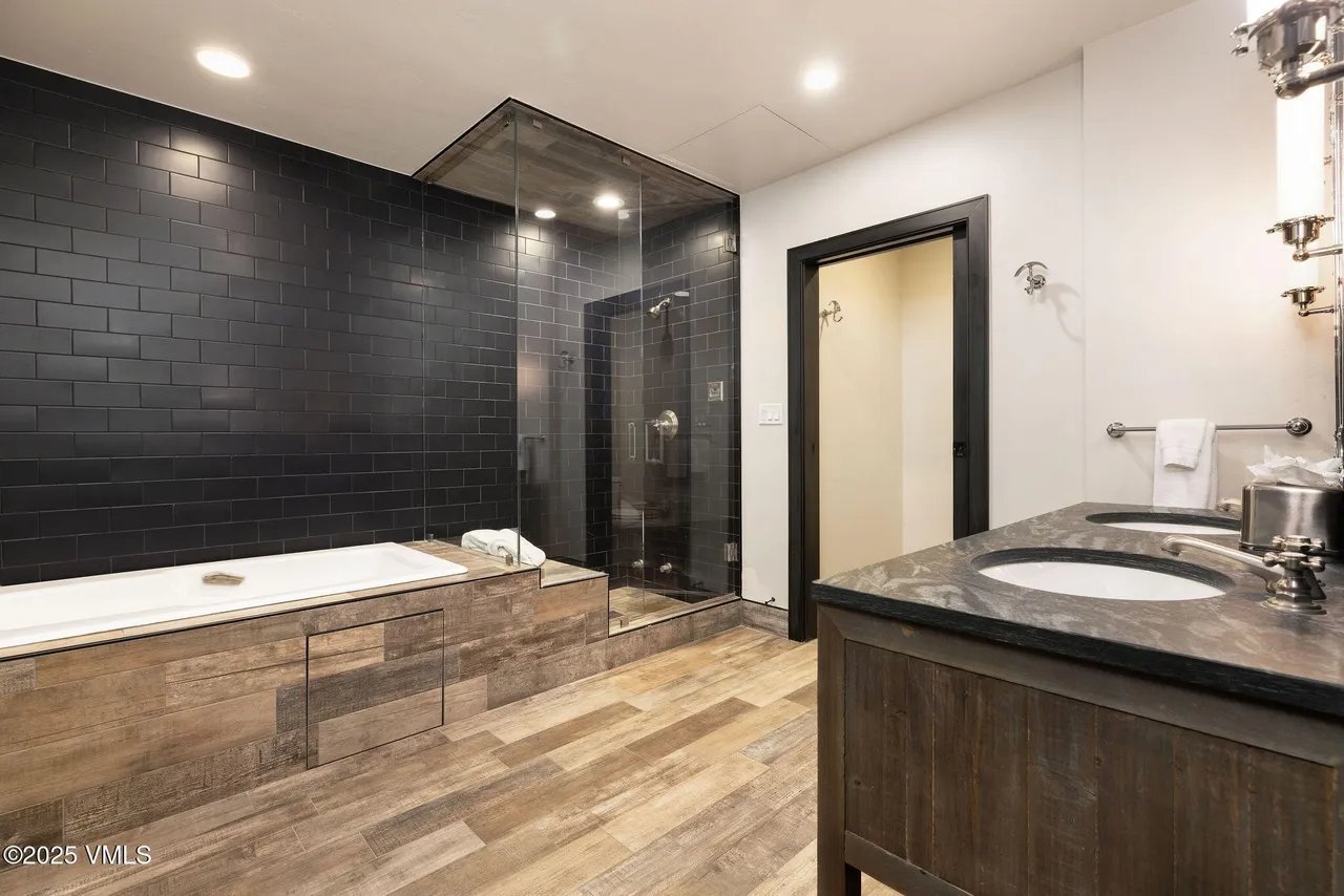 This is a contemporary primary bathroom featuring a dark tile shower and tub surround. The vanity has dual sinks with a dark countertop. The floors are wood-look tile, creating a spa-like ambiance.