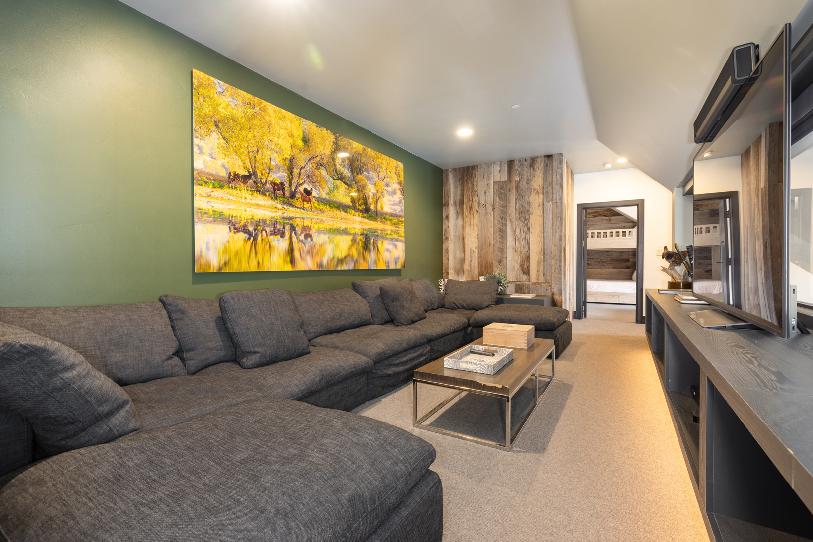 This is a cozy living room featuring a large sectional sofa with generous cushions. The room is decorated with an oversized landscape painting and has unique wood paneling. A modern coffee table and built-in entertainment unit add to the room's functionality and appeal for relaxation.