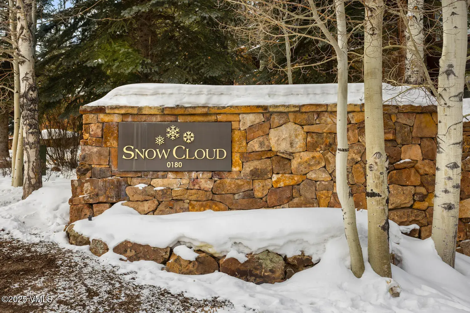The image showcases the stone entryway sign for "Snow Cloud 0180," set against a backdrop of birch trees and snow-covered ground. The sign itself is made of rough-cut stones, topped with a darker plaque displaying the property name and number. The overall impression is of a mountain property with a rustic yet elegant welcome.