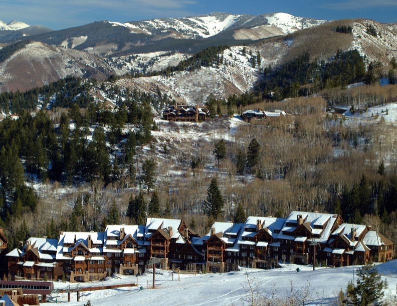 This is an exterior front view of a mountain resort property with multi-story townhouses or condos. The buildings feature a rustic design with wood and stone elements, nestled in a snowy landscape. Mountains covered with snow in the background add to the scenic appeal of the location, suggesting a ski-in/ski-out experience.