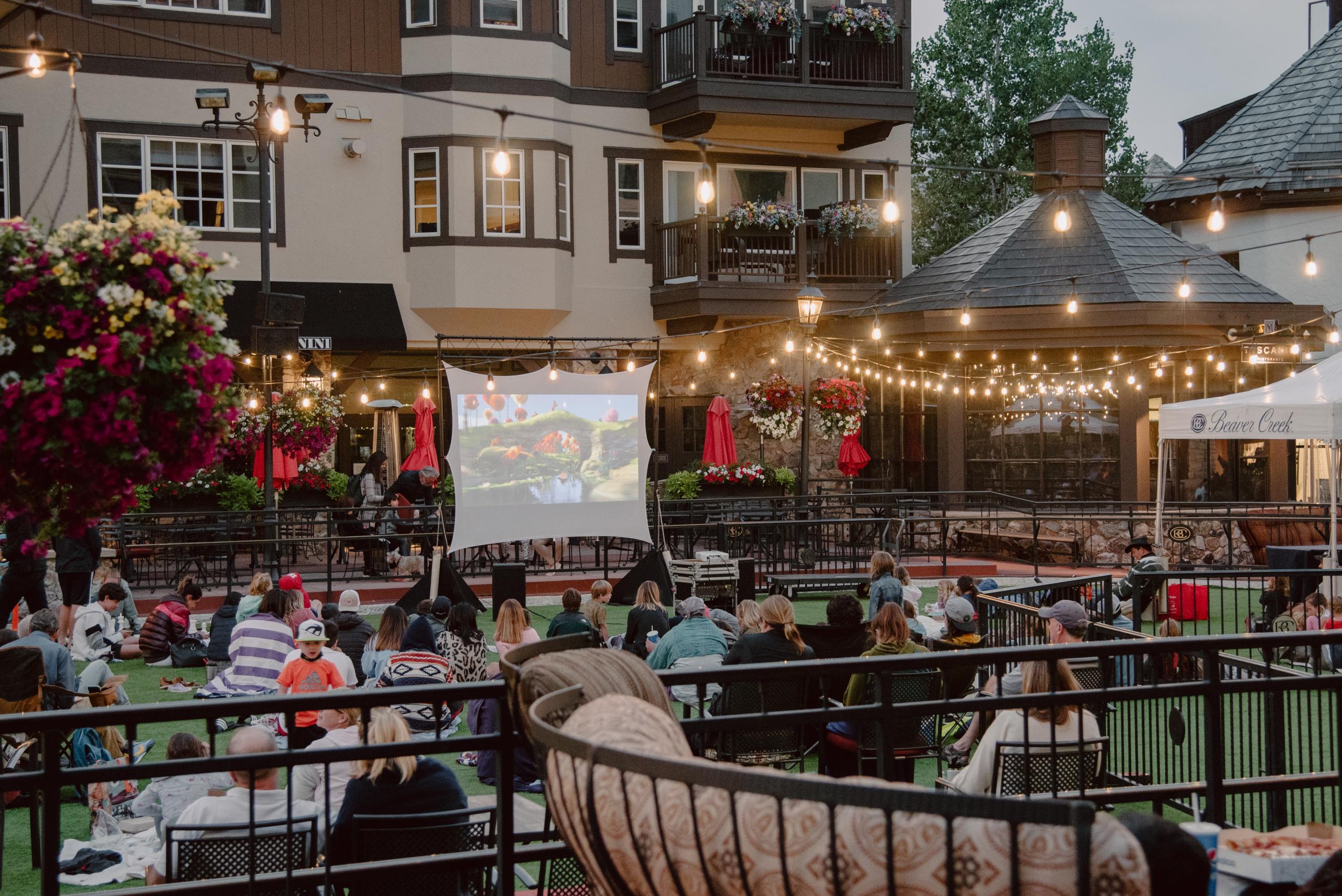 This image captures an outdoor movie night in a community space, likely a common area within a residential development. A large screen displays a film to a seated audience on an artificial lawn, with string lights adding an inviting ambiance. The setup suggests a family-friendly and community-oriented atmosphere, enhancing the appeal for potential residents.
