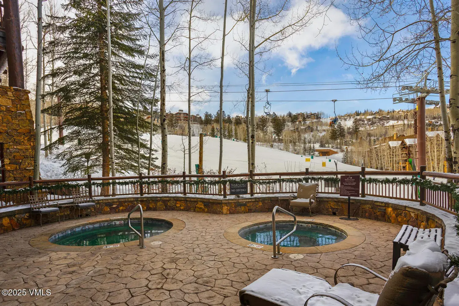 This image showcases a luxury outdoor spa area at a mountain resort. Two hot tubs are visible, surrounded by stone paving, with a decorative wooden fence and winter foliage. The background features a snowy hillside and a ski lift, emphasizing the property's proximity to skiing amenities.