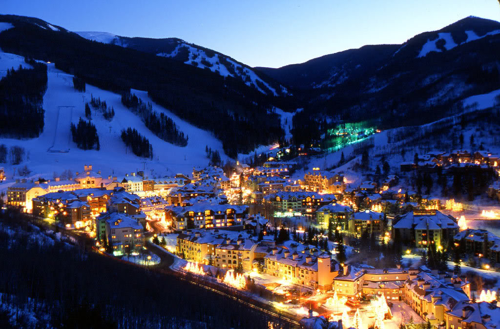 This aerial view showcases a picturesque ski resort town at night. The snow-covered mountains provide a stunning backdrop to the illuminated chalets and buildings below. The scene evokes a sense of luxury and relaxation, ideal for a winter getaway.