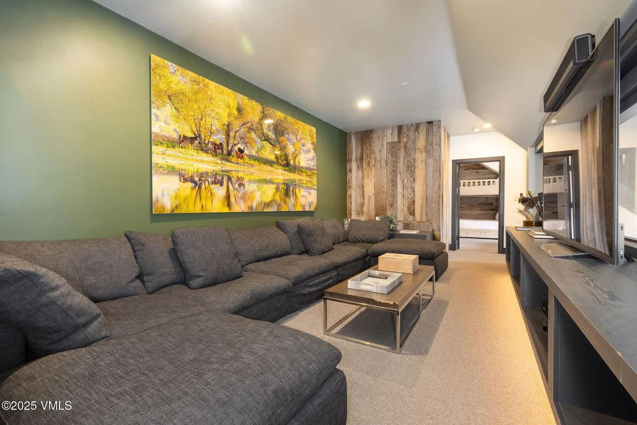 This inviting living room features a large, comfortable gray sectional sofa, perfect for relaxation and entertaining. A striking landscape artwork adorns the wall, complemented by rustic wood paneling. The room's design is both cozy and stylish, offering a blend of modern comfort and natural elements.