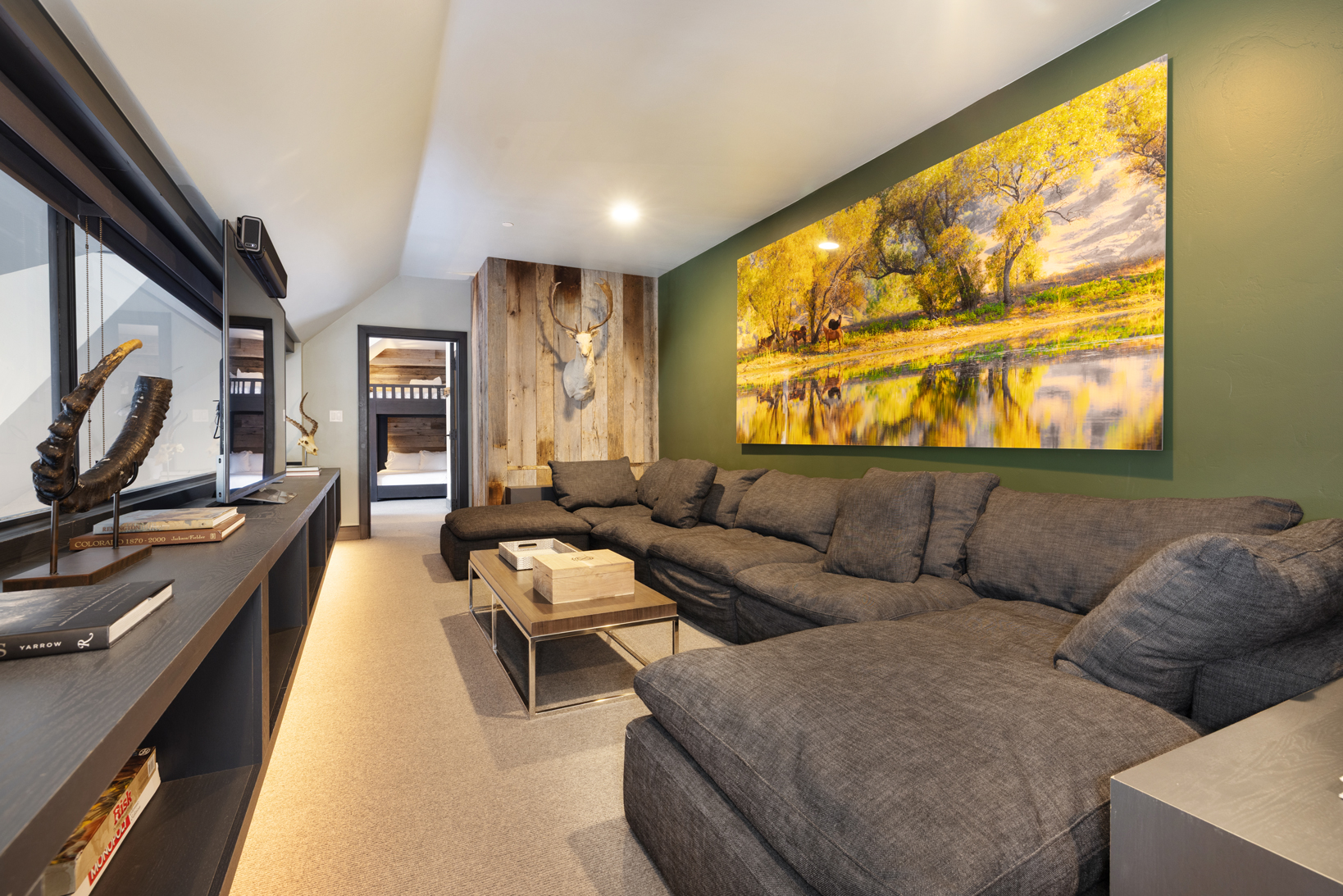 This living room features a large, comfortable sectional sofa, an eye-catching nature-themed artwork, and a rustic accent wall with a mounted deer head. A modern media console with display shelving adds a touch of contemporary style, creating a cozy yet sophisticated atmosphere. The room appears well-lit and inviting, perfect for relaxing or entertaining.