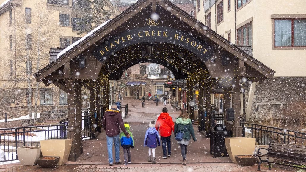 This image depicts a family walking through an entryway marked by Beaver Creek Resort signage, set against a backdrop of snowy weather and buildings. The entryway is decorated with string lights, providing a welcoming ambiance, while the surrounding architecture suggests a luxury resort environment, making it an appealing destination. The presence of snow enhances the winter vacation appeal.