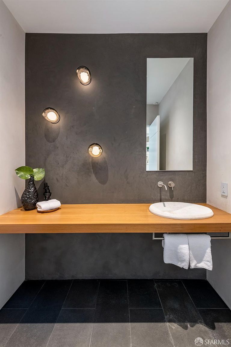 This modern powder room features a striking dark, textured accent wall illuminated by three circular wall-mounted light fixtures. A floating light-wood vanity provides a warm contrast, topped with a circular stone vessel sink and wall-mounted faucets. A rectangular mirror is mounted above the sink, while a towel bar below holds white linens, all set against dark floor tiles.