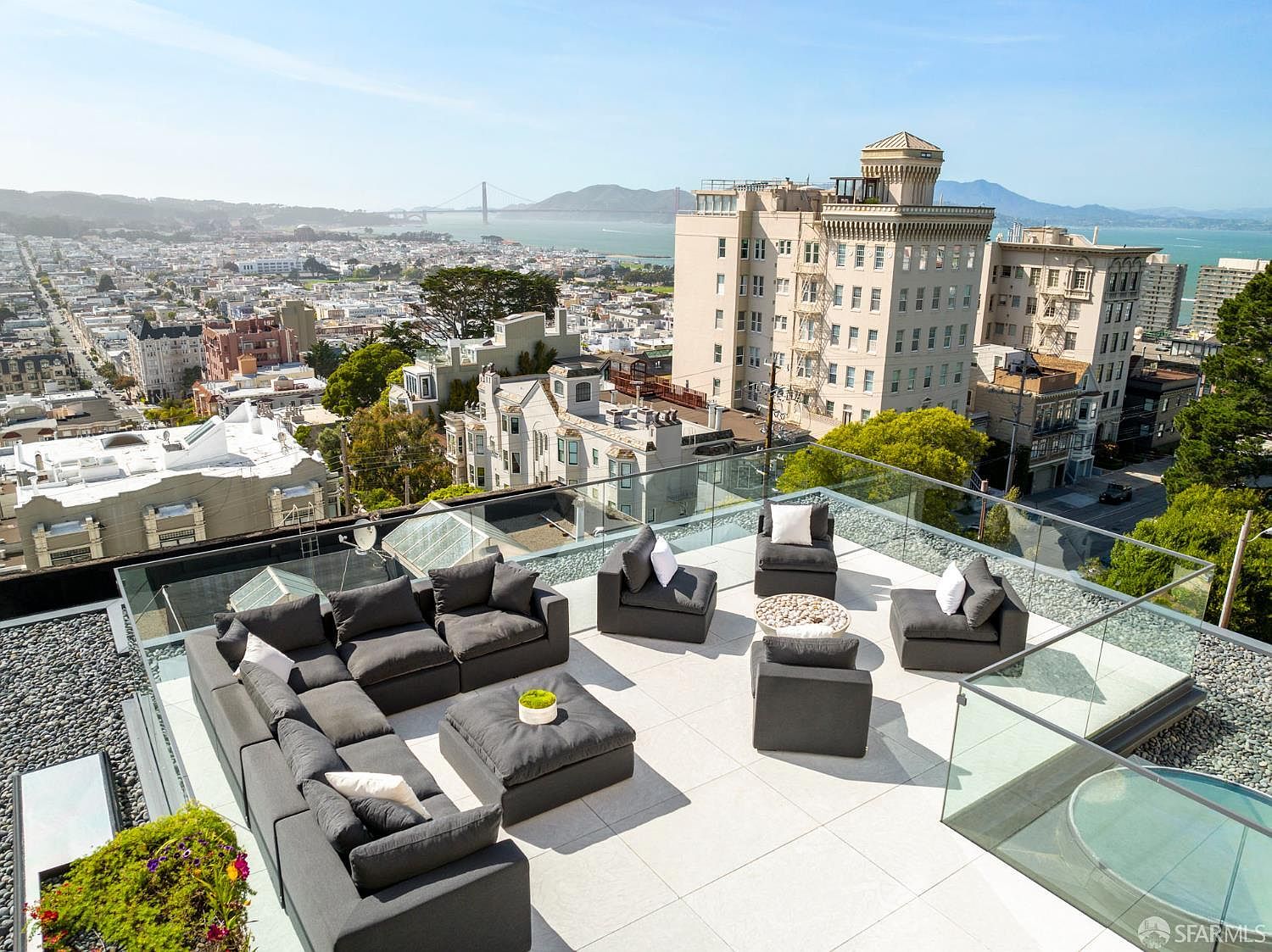 This expansive rooftop terrace offers a luxurious outdoor living space with a panoramic view of the San Francisco skyline and the Golden Gate Bridge. The area is furnished with a large, modular dark gray sectional sofa, a matching ottoman, and several individual lounge chairs arranged around a central fire pit. A sleek glass railing surrounds the perimeter, ensuring unobstructed views while maintaining a modern, sophisticated aesthetic.