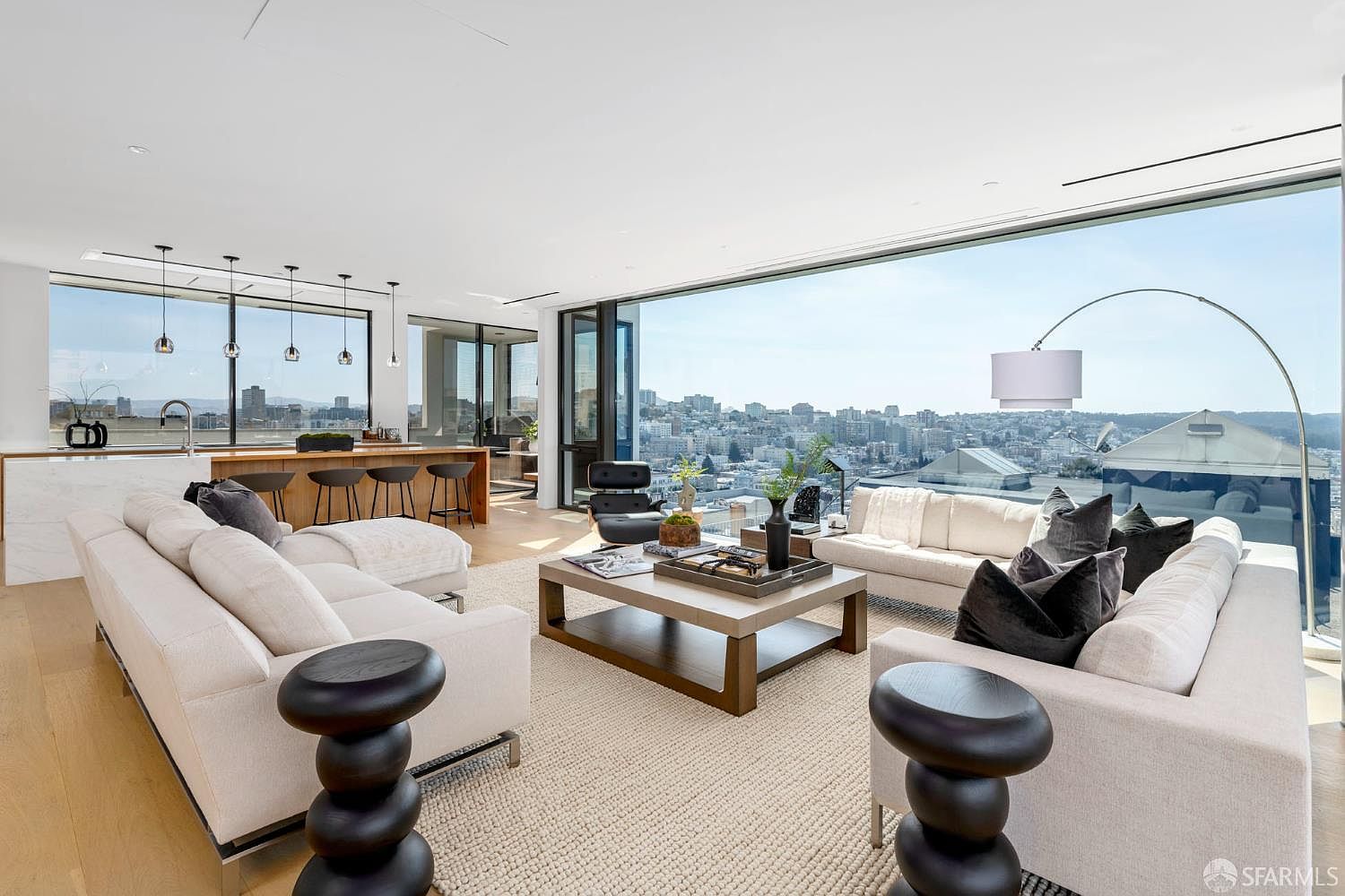 This expansive, modern living room features a bright, open-concept layout with light wood flooring and a neutral color palette. The space is defined by floor-to-ceiling windows that provide a panoramic city view, complemented by two large sofas, a central coffee table, and sculptural side tables. A kitchen area with a breakfast bar and pendant lighting is visible in the background, creating a seamless transition between living and dining spaces.
