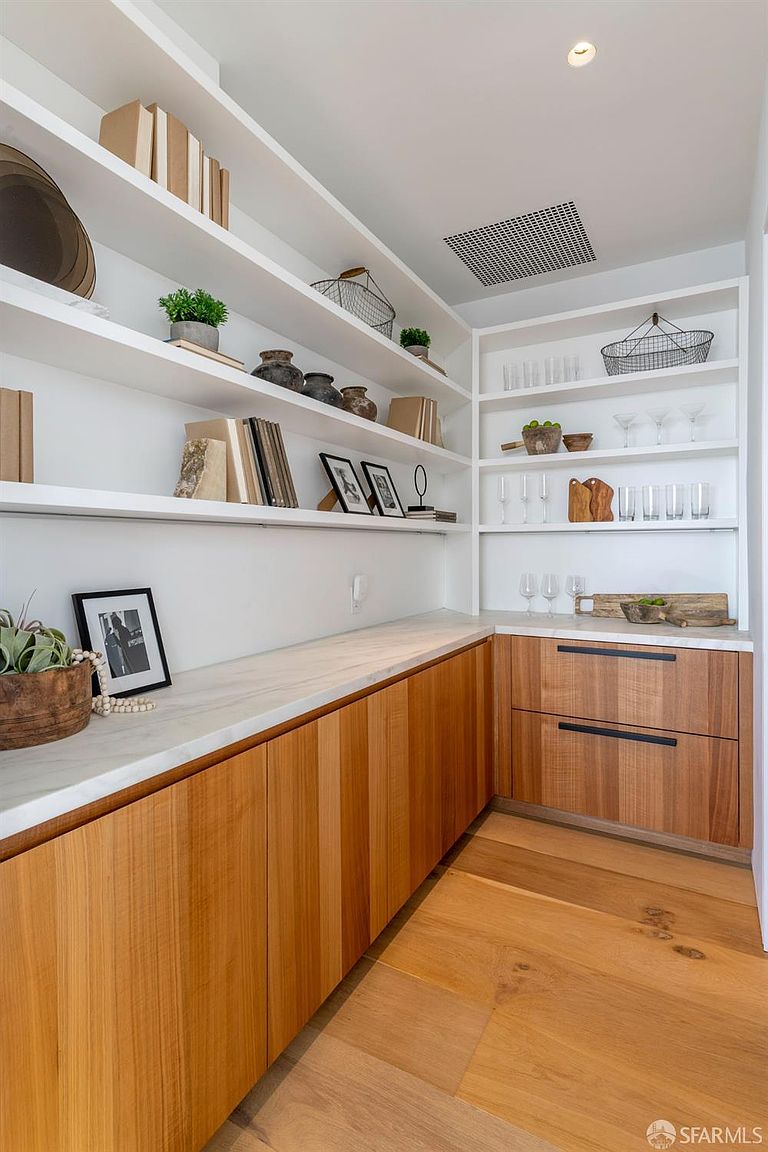This organized pantry or butler's pantry features light wood cabinetry with clean lines and a smooth stone countertop. Open shelving lines the walls, displaying a curated collection of glassware, decorative pottery, and books, creating a bright and functional storage space. The perspective is a wide-angle shot from the corner, highlighting the seamless integration of the cabinetry and the warm, natural wood flooring.