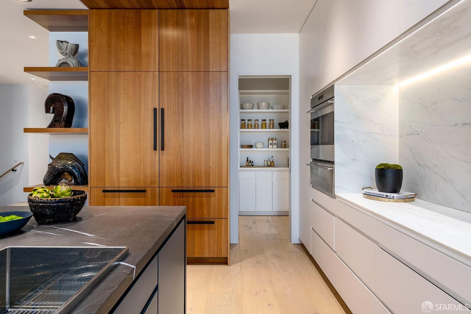 This modern kitchen features a striking contrast between warm wood-paneled cabinetry and sleek white surfaces. A large island with a dark stone countertop sits in the foreground, while a walk-in pantry is visible through a central doorway. The space is accented by open shelving displaying decorative objects and integrated appliances, creating a sophisticated and functional culinary environment.
