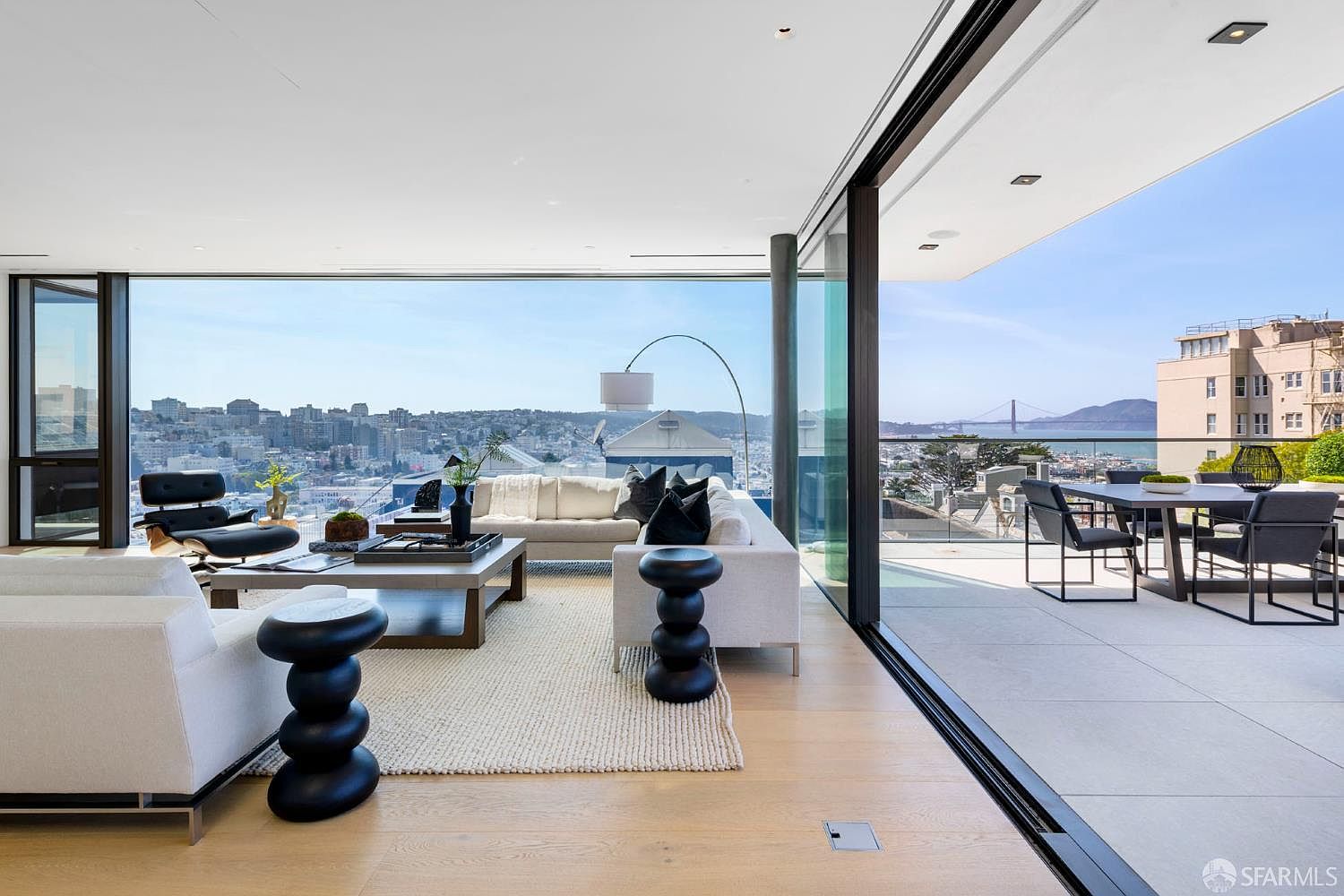 This sophisticated living room features an open-concept layout that seamlessly transitions to an expansive outdoor terrace through large, retractable glass walls. The interior is furnished with a plush white sofa, a classic lounge chair, and unique sculptural side tables, all set against a backdrop of panoramic city and bay views. The space is defined by its clean lines, light wood flooring, and a bright, airy atmosphere that emphasizes the connection between indoor and outdoor living.