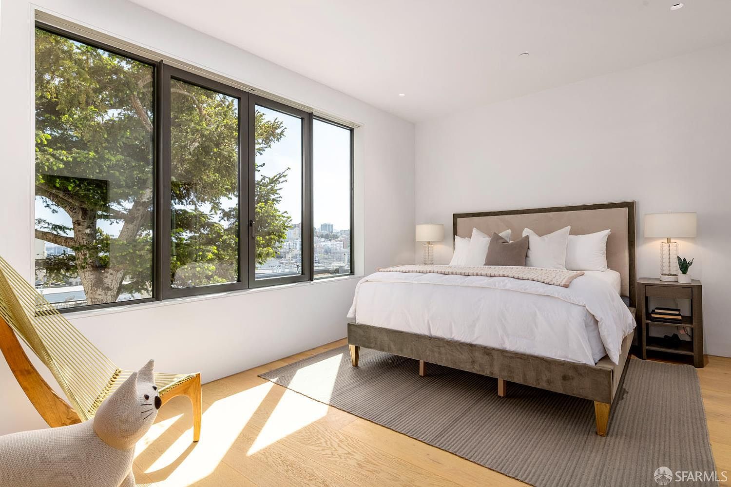 This bright and airy primary bedroom features a large bed with a neutral-toned upholstered headboard, centered against a clean white wall. A set of large windows provides ample natural light and a view of the surrounding neighborhood and trees, while light wood flooring and a textured area rug add warmth to the space. A whimsical cat-shaped chair sits in the foreground, and matching bedside tables with lamps complete the minimalist, contemporary aesthetic.