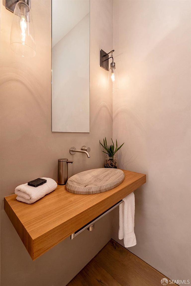 This minimalist powder room features a floating light wood vanity topped with a unique, oval-shaped stone vessel sink. A wall-mounted faucet and a sleek, vertical mirror are centered above the vanity, flanked by two modern wall sconces that provide warm lighting. The walls are finished in a smooth, neutral-toned plaster, creating a serene and contemporary atmosphere.