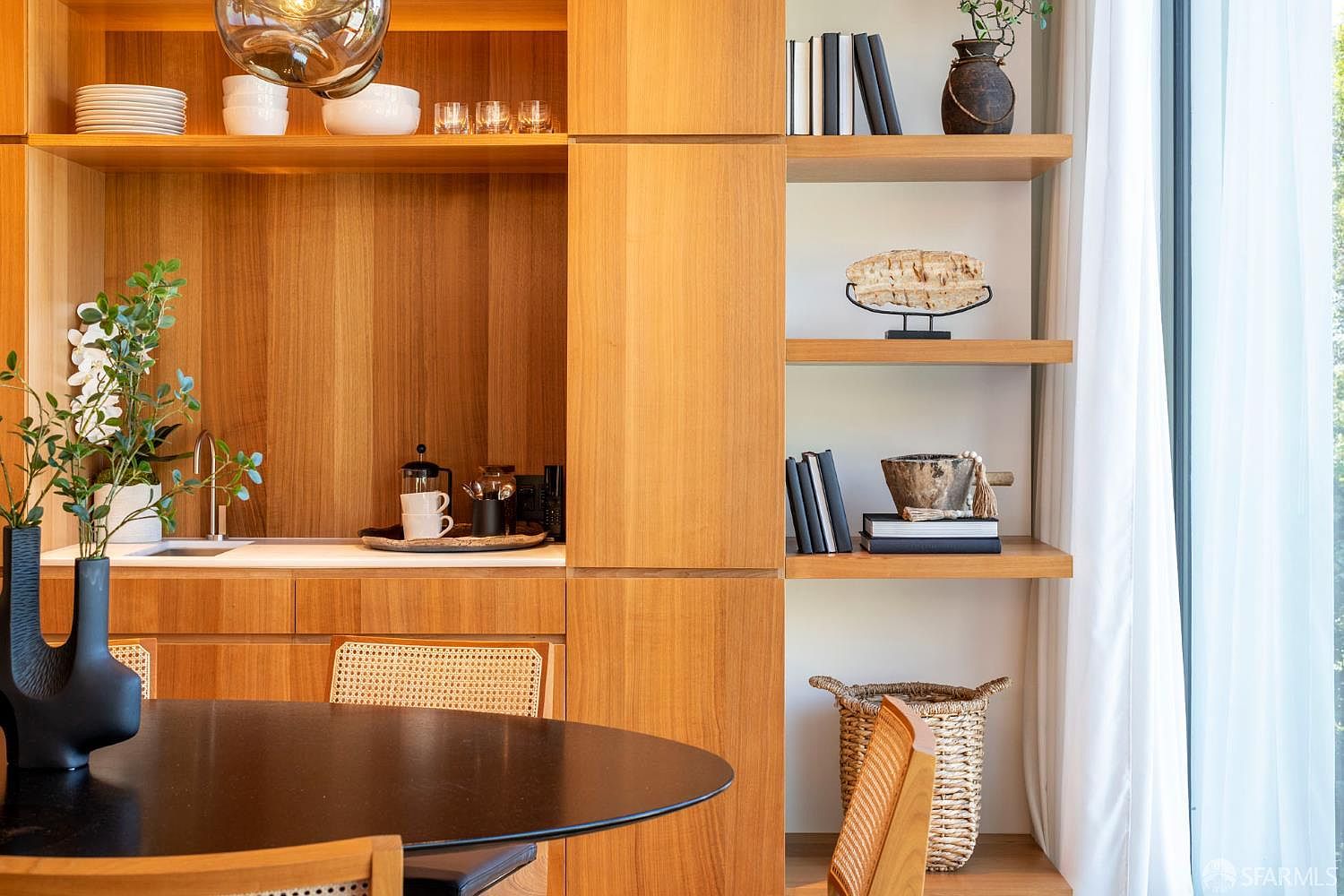 This dining area features a sleek, round black table paired with light wood chairs that have woven cane detailing. A custom-built light wood cabinetry unit serves as a wet bar, complete with an integrated sink, open shelving for display, and a dedicated space for coffee service. The composition is framed by a tall window with soft white curtains, creating a warm, sophisticated, and functional space.