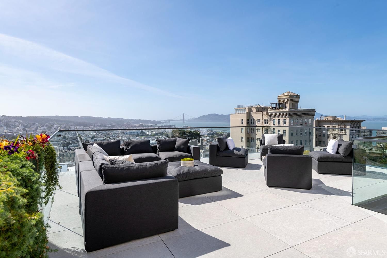 This expansive rooftop terrace offers a sophisticated outdoor living space with a modular dark gray sectional sofa and matching ottoman. The area is framed by a sleek glass railing that provides an unobstructed, panoramic view of the city skyline and the Golden Gate Bridge in the distance. A vibrant planter box filled with colorful flowers adds a touch of natural beauty to the modern, minimalist aesthetic of the deck.