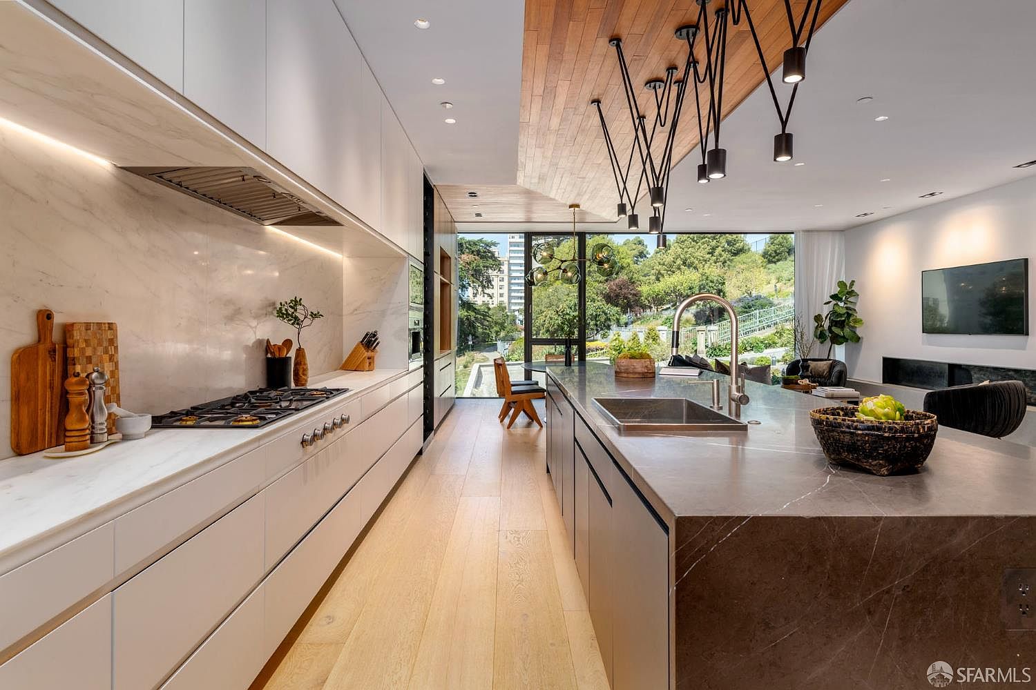 This modern, open-concept kitchen features sleek white cabinetry paired with a large, dark stone-topped island and light wood flooring. A unique, multi-armed pendant light fixture hangs from a wood-paneled ceiling, drawing the eye toward the large windows that provide a view of the lush greenery outside. The space is designed with a minimalist aesthetic, emphasizing clean lines and a seamless transition between the culinary area and the adjacent living space.