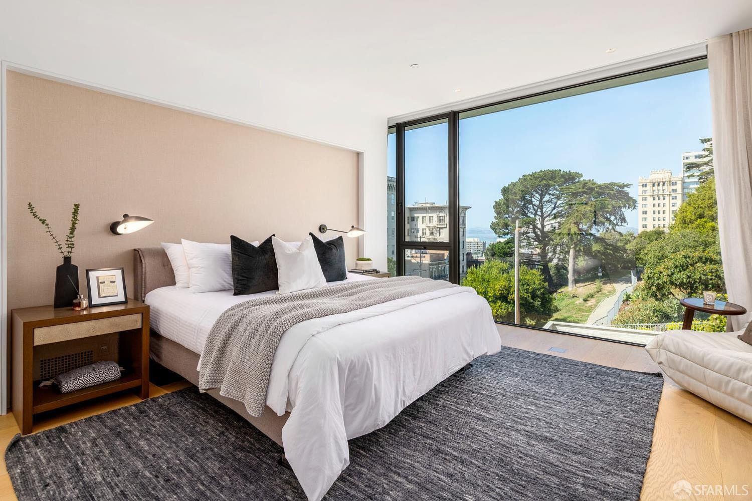 This modern primary bedroom features a neutral color palette with a textured accent wall behind the bed. A large, floor-to-ceiling window provides an expansive view of the surrounding trees and city buildings, filling the space with natural light. The room is furnished with a plush bed, a dark wood nightstand, and a textured area rug, creating a serene and sophisticated atmosphere.
