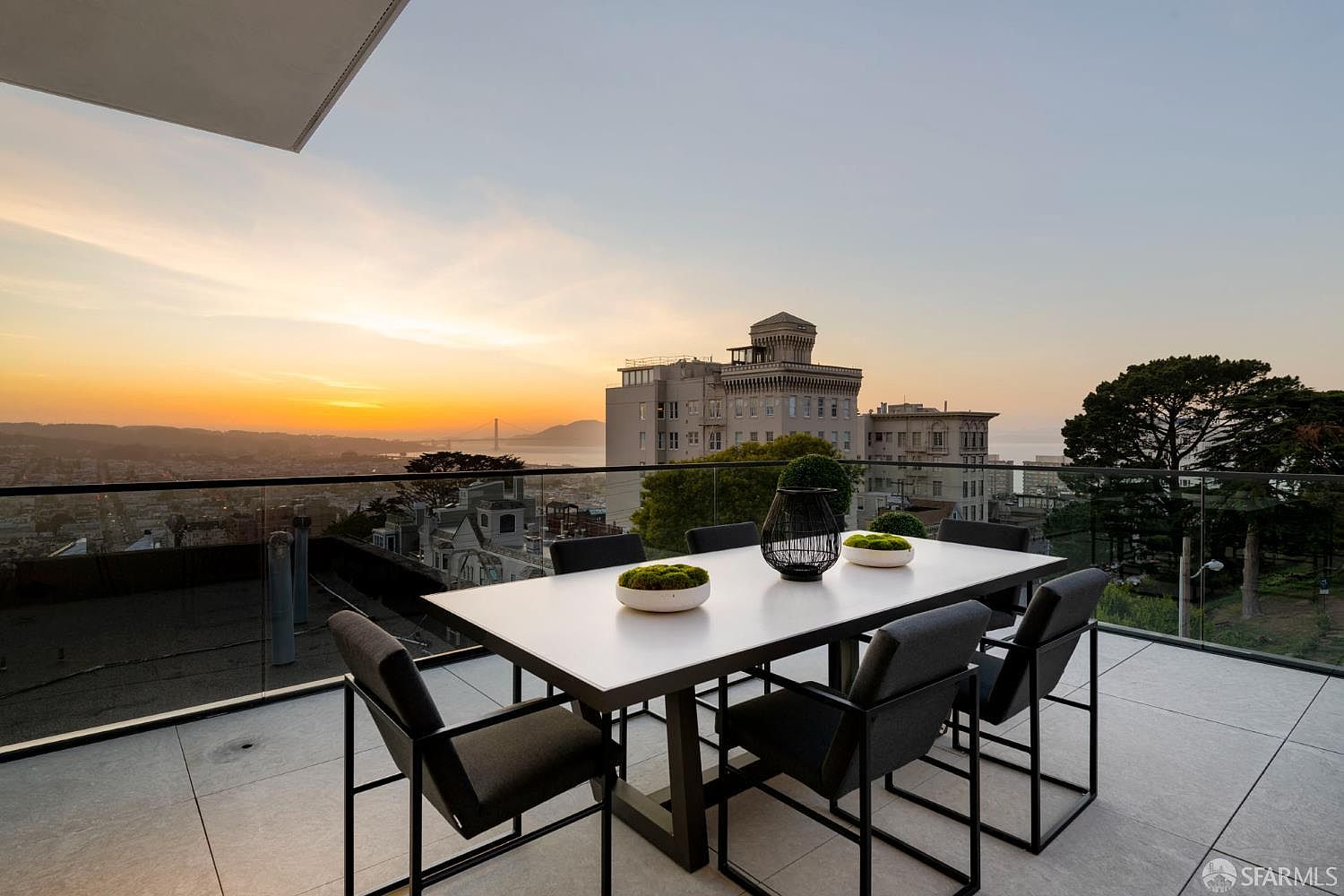 This high-rise balcony features a sleek, modern dining set with a white rectangular table and six dark-upholstered chairs, perfect for outdoor entertaining. The space is enclosed by a frameless glass railing that provides an unobstructed, panoramic view of the city skyline and the Golden Gate Bridge at sunset. The clean, minimalist aesthetic is complemented by the tiled flooring and simple decorative greenery on the table, creating an elegant and serene atmosphere.