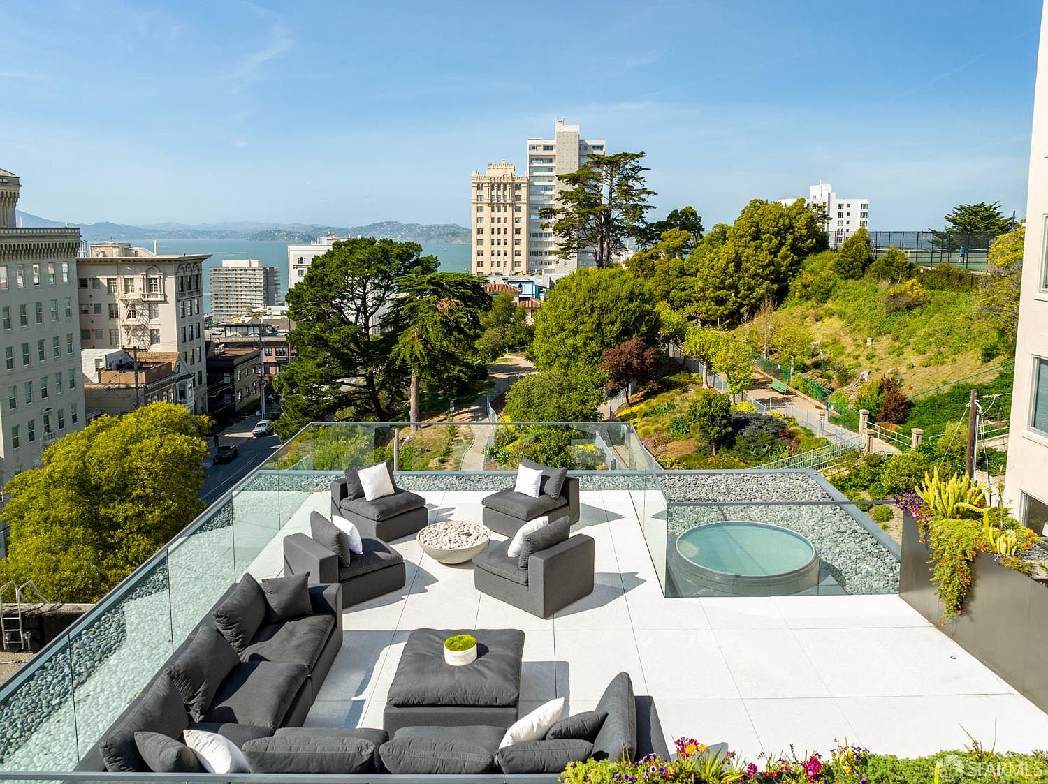 This expansive rooftop terrace offers a luxurious outdoor living space with panoramic city and bay views. The area is furnished with a large modular sectional sofa, several lounge chairs, and a central fire pit, all set against a backdrop of lush greenery and urban architecture. A circular spa is integrated into the deck, which is enclosed by a sleek glass railing that maintains an unobstructed view of the surrounding landscape.