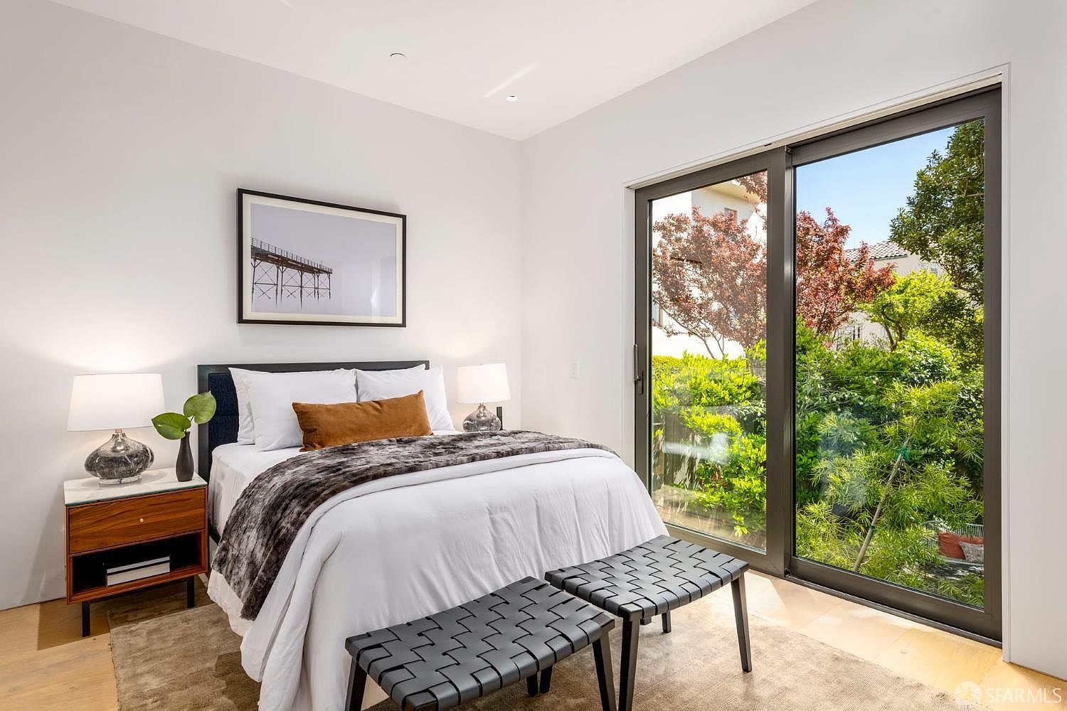 This modern bedroom features a queen-sized bed with a dark headboard, crisp white bedding, and a textured throw blanket. Two black woven benches sit at the foot of the bed, while a mid-century inspired nightstand with a lamp provides bedside storage. A large sliding glass door offers a bright, expansive view of the lush green foliage outside, creating a serene and airy atmosphere.