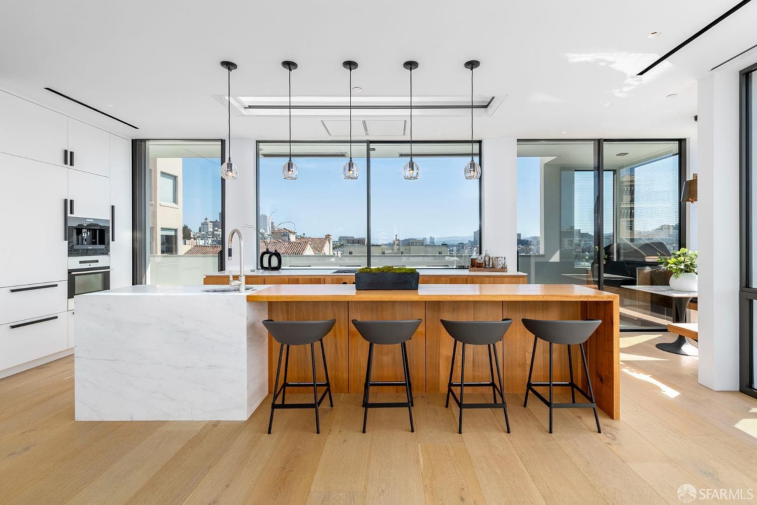 This modern, open-concept kitchen features a large island with a stone waterfall edge and light wood cabinetry, complemented by four dark bar stools. The space is illuminated by five minimalist pendant lights hanging above the island, set against a backdrop of expansive windows that provide a clear view of the city skyline. The clean, minimalist aesthetic is enhanced by light wood flooring and integrated appliances, creating a bright and sophisticated atmosphere.