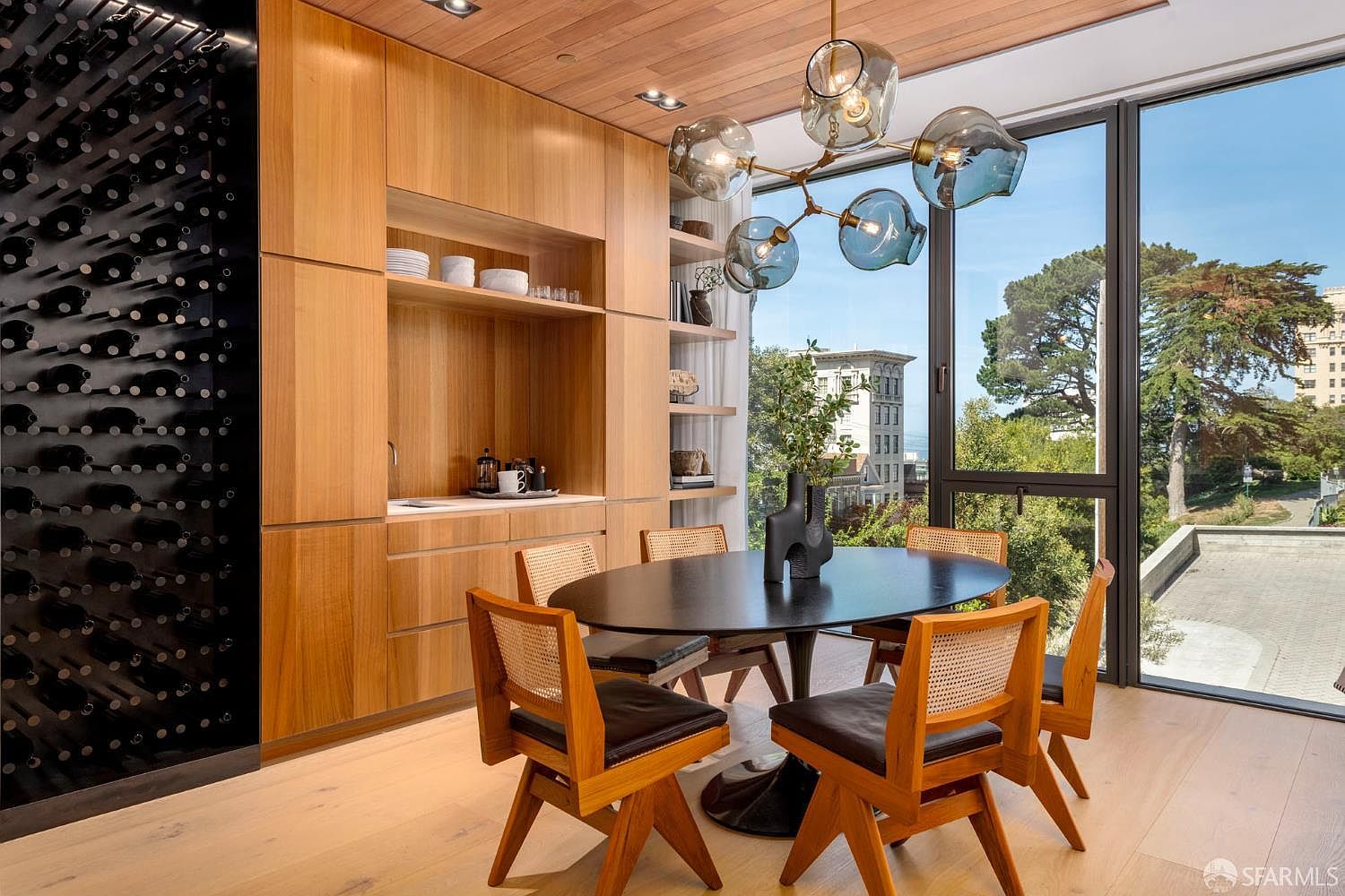This sophisticated dining area features a dark oval table surrounded by chairs with woven cane backs and dark leather seats. A built-in light wood cabinetry unit includes a wet bar and open shelving, complemented by a striking black wine storage wall to the left. Large windows provide an expansive view of the outdoors, while a modern, multi-globe chandelier serves as a focal point above the table.
