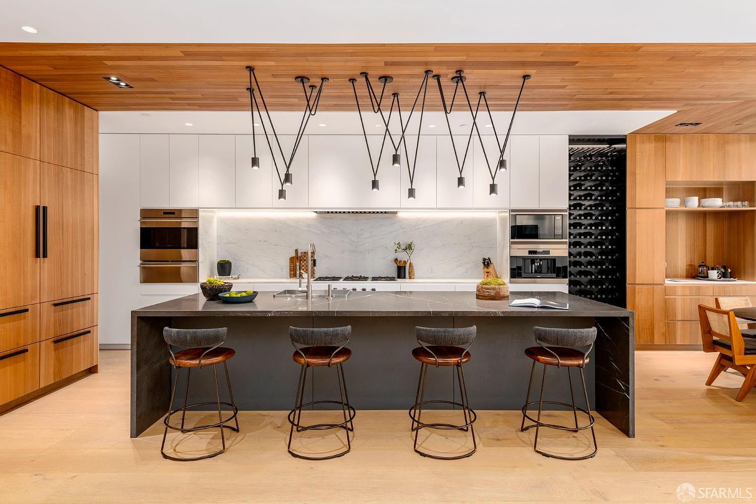 This modern, high-end kitchen features a large central island with a dark stone countertop and four bar stools, set against a backdrop of white cabinetry and light wood paneling. The space is defined by a striking geometric pendant lighting arrangement suspended from a warm wood-paneled ceiling, creating a sophisticated and cohesive aesthetic. A built-in wine storage unit and integrated appliances contribute to the sleek, minimalist design, while the open layout provides a seamless transition to the adjacent dining area.
