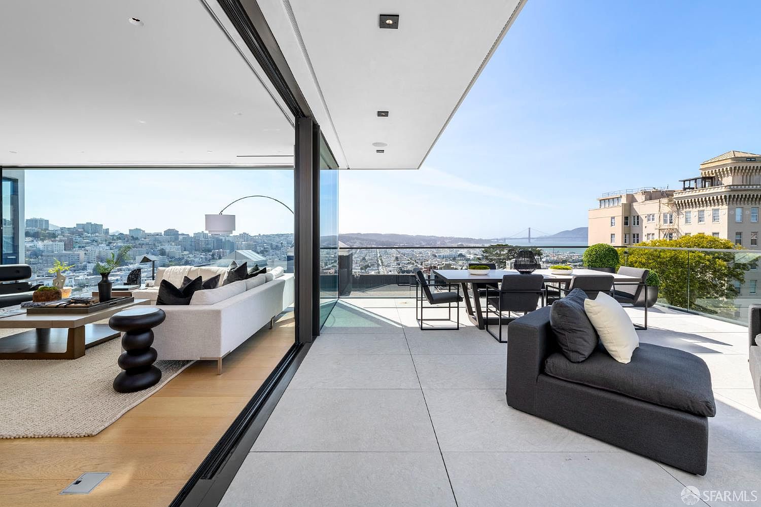 This expansive, modern balcony offers a seamless transition from the indoor living area through large, retractable glass walls. The outdoor space features a dining set and a comfortable lounge chair, all set against a panoramic view of the city skyline and the Golden Gate Bridge. The clean lines and neutral color palette emphasize a sophisticated, high-end urban lifestyle.
