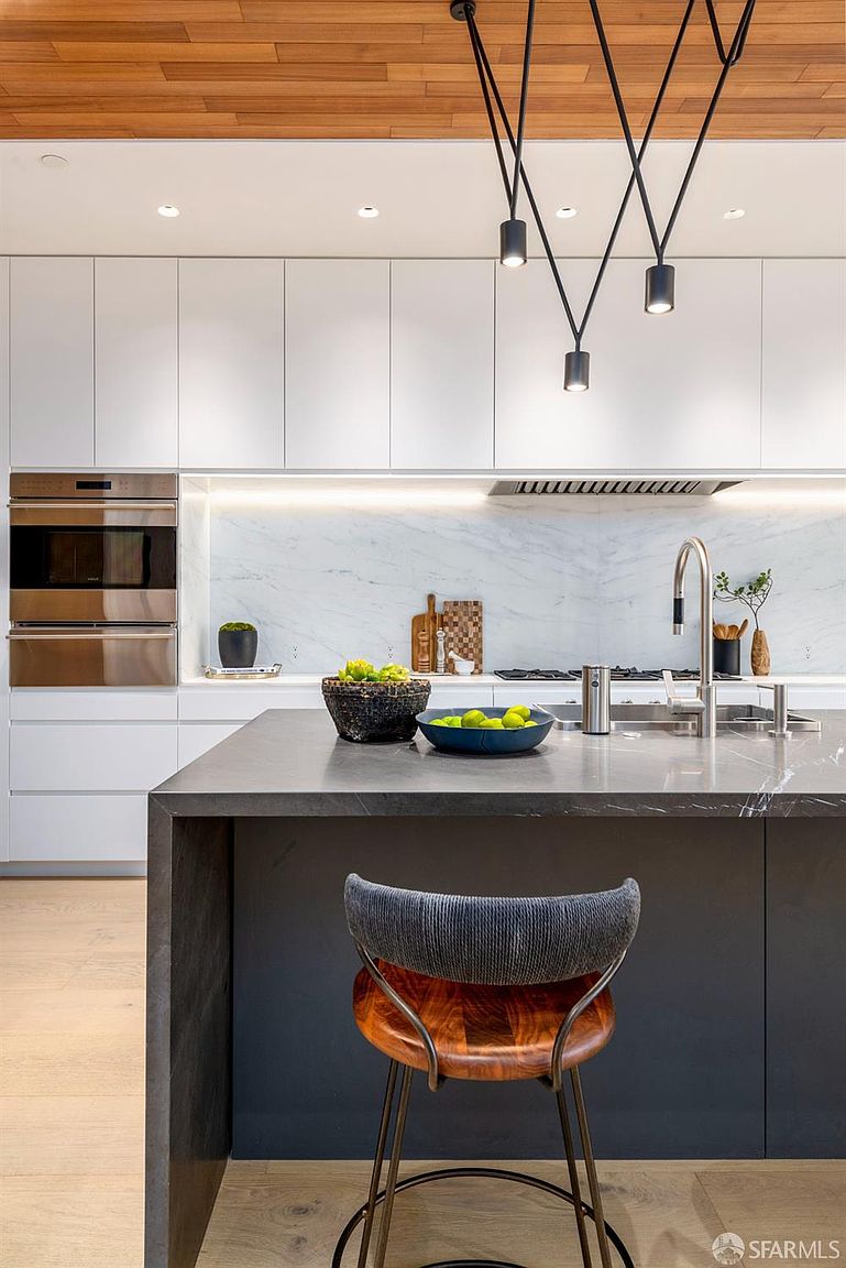 This modern kitchen features a sleek, minimalist design with white cabinetry, a dark stone-topped island, and a light wood ceiling. A striking geometric pendant light fixture hangs above the island, which is complemented by a marble-look backsplash and integrated stainless steel appliances. The perspective is centered on the island, highlighting the clean lines and high-end finishes of the space.