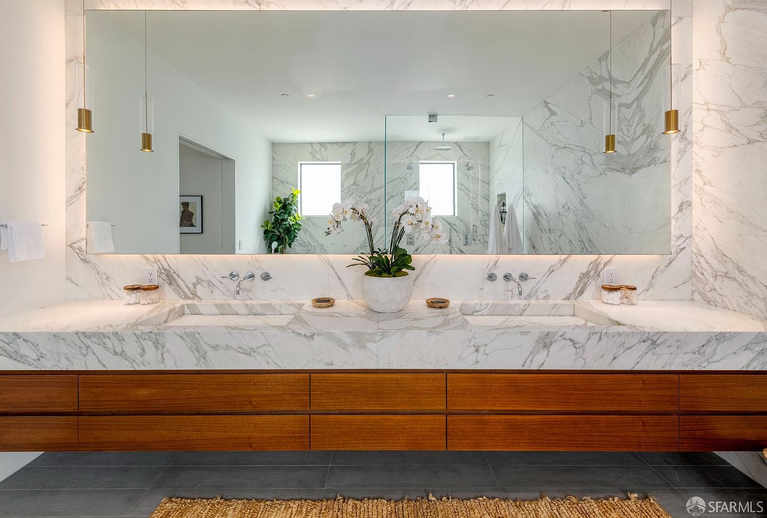 This primary bathroom features a sleek, double-vanity setup with a long, stone-topped counter and warm-toned wood cabinetry. A large, wall-spanning mirror reflects the space, including a central orchid arrangement and modern pendant lighting fixtures. The room is finished with light-veined stone wall cladding and dark, large-format floor tiles, creating a sophisticated and contemporary aesthetic.