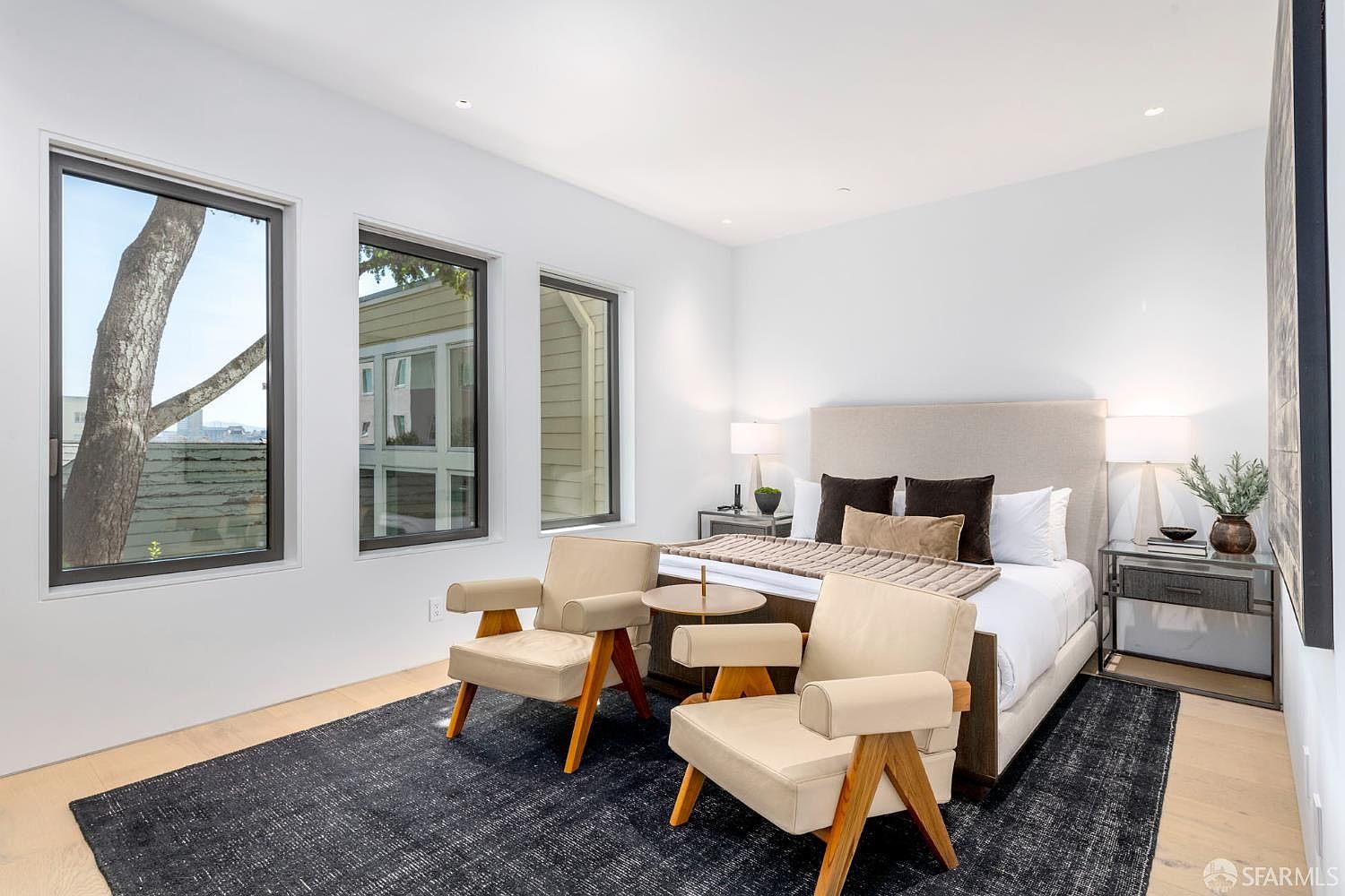This bright and airy primary bedroom features a neutral color palette with light wood flooring and a large, dark area rug. The room is furnished with a plush bed, two modern armchairs, and matching bedside tables with lamps, creating a sophisticated and tranquil atmosphere. Three large windows along the wall provide ample natural light and a view of the exterior surroundings.