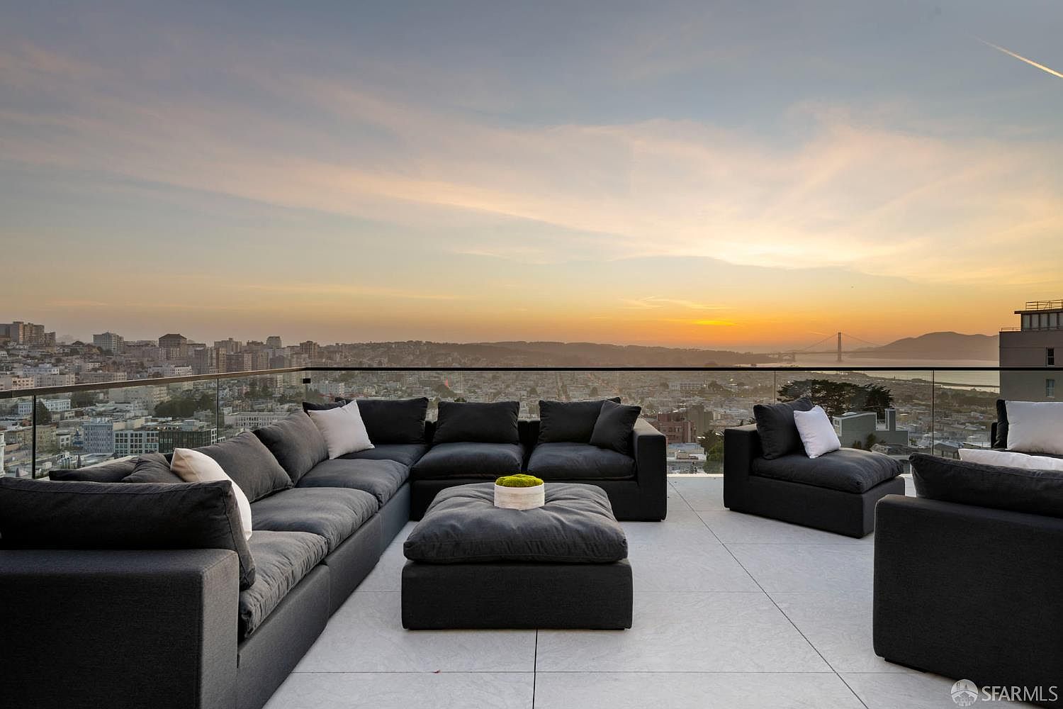 This expansive rooftop terrace offers a luxurious outdoor living space with a large, modular dark gray sectional sofa and a matching ottoman centered around a small decorative plant. The area is framed by a sleek, transparent glass railing that provides an unobstructed, panoramic view of the city skyline and the Golden Gate Bridge at sunset. The scene captures a sophisticated urban atmosphere, perfect for entertaining against a dramatic, colorful evening sky.