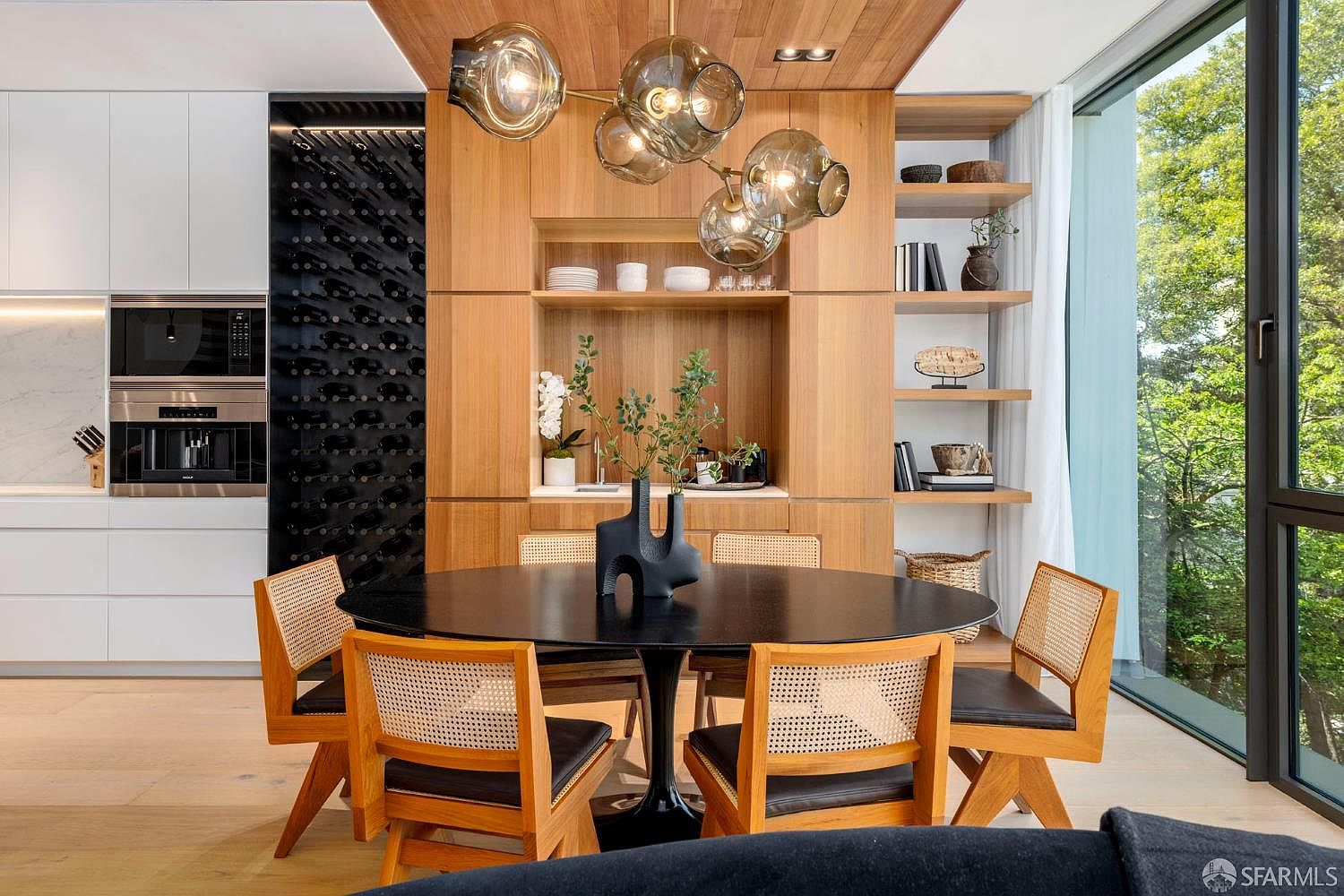 This sophisticated dining area features a round black table surrounded by four chairs with light wood frames and woven cane backs. The space is anchored by a warm wood-paneled wall that incorporates open shelving and a built-in wine storage rack, while a striking multi-globe pendant light hangs above the table. Large windows to the right provide an expansive view of lush greenery, creating a seamless connection between the modern interior and the outdoors.