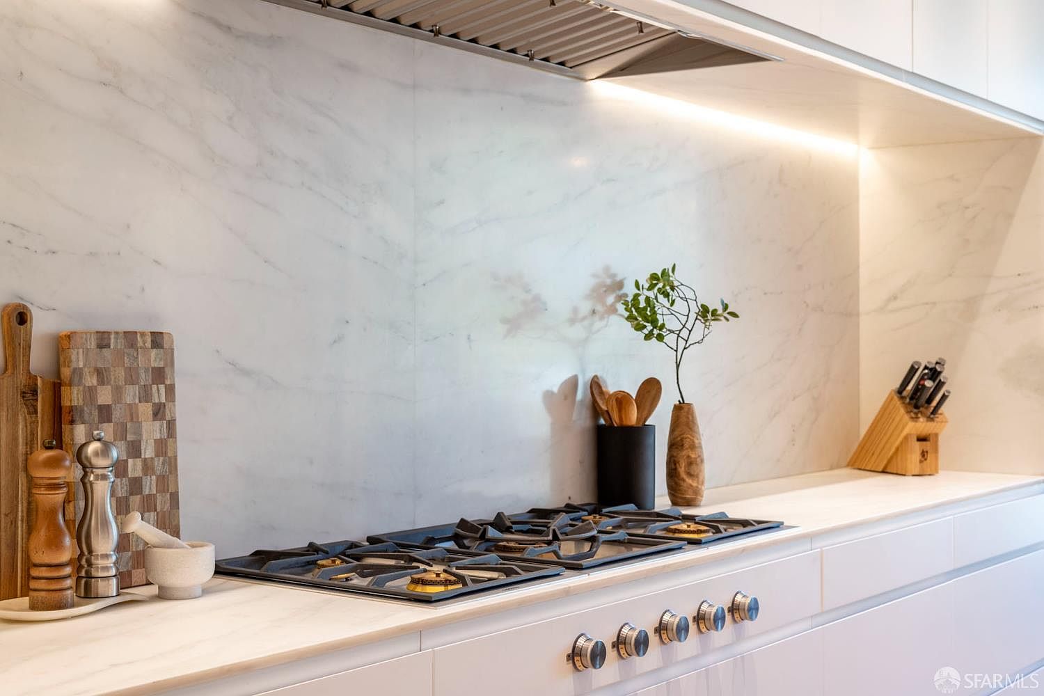 This sleek, modern kitchen features a gas cooktop set into a light-toned stone countertop with a matching full-height stone backsplash. The space is accented by a minimalist range hood, a wooden knife block, and a decorative vase with greenery, creating a clean and sophisticated aesthetic. The perspective is a close-up, eye-level shot that highlights the high-end finishes and organized culinary workspace.