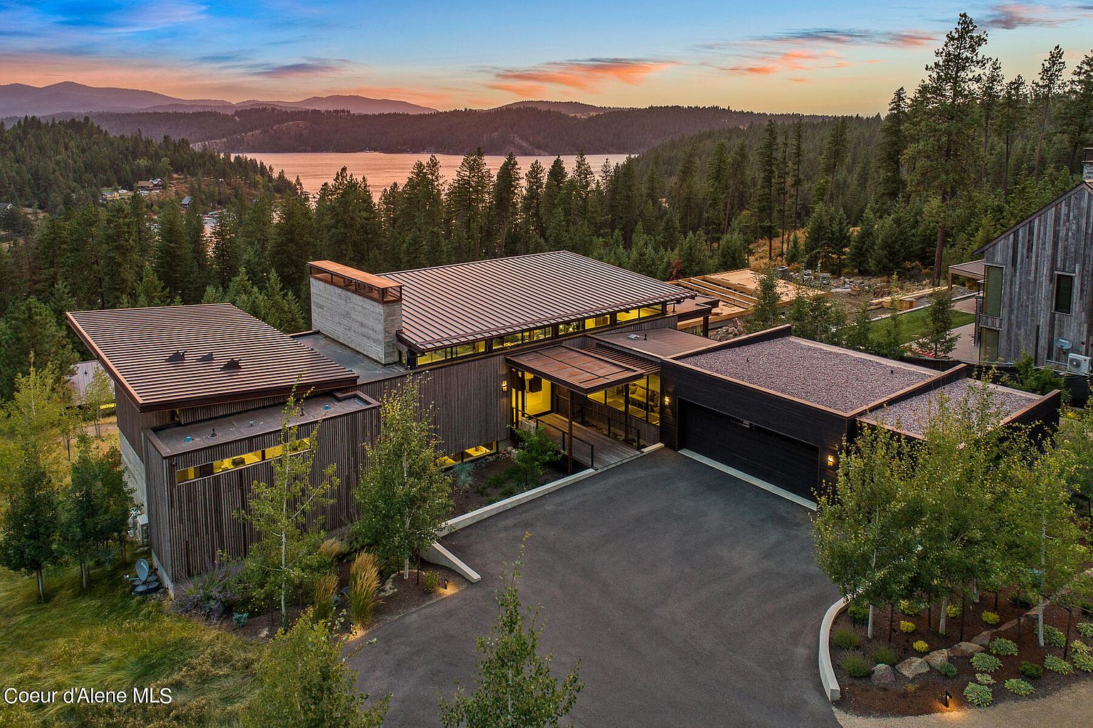This aerial view showcases a modern, multi-structure home nestled amidst a lush forest setting with a lake and mountains in the background. The house features a combination of wood and stone elements, flat and sloped roofs, and a spacious driveway leading to a garage. The landscape is well-maintained, enhancing the property's curb appeal and highlighting its secluded location.
