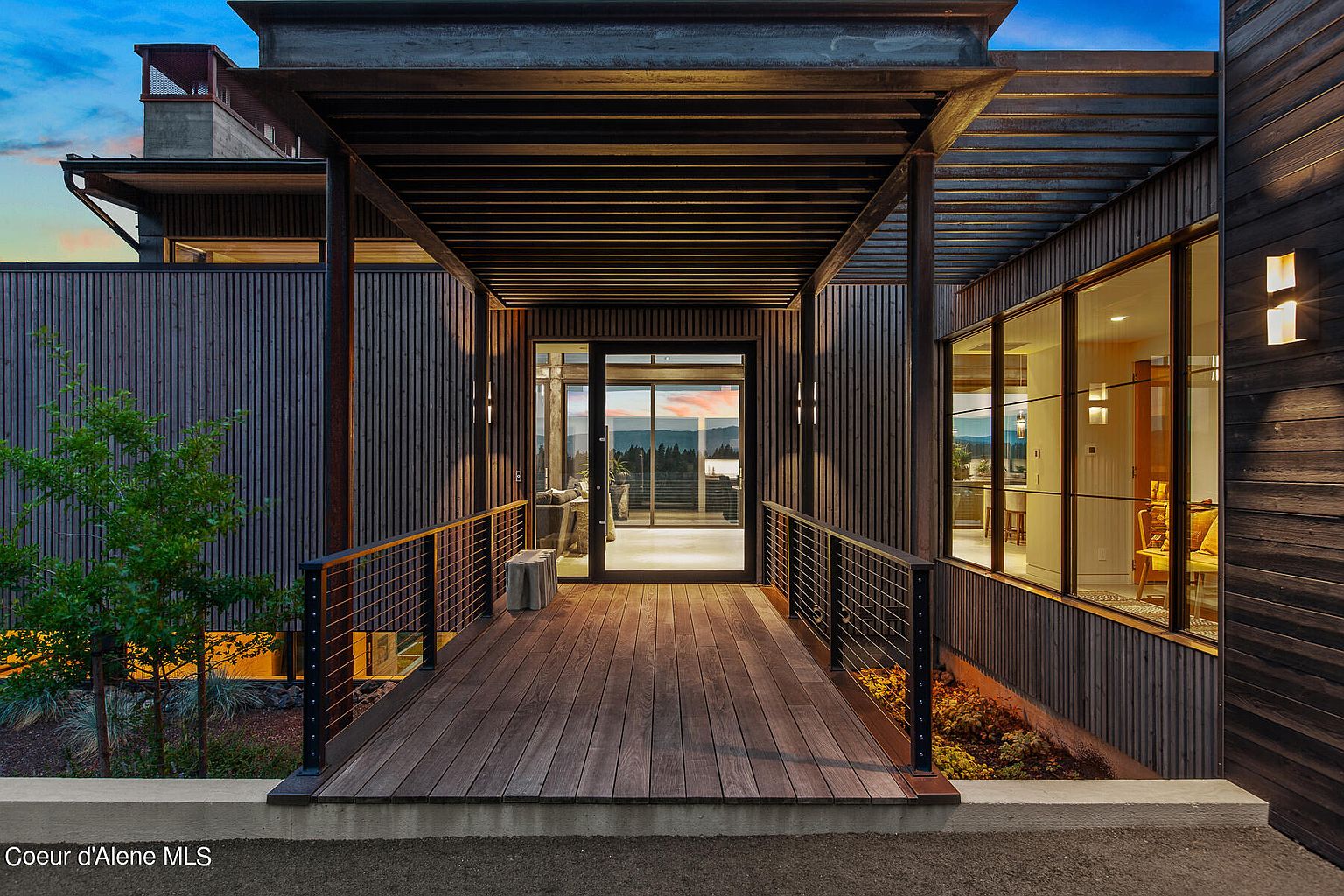 This image showcases a modern home's entryway, featuring a wooden walkway leading to a glass-door entrance. The structure is defined by dark, vertical wood siding and a striking metal pergola overhead. The design emphasizes clean lines and natural materials, creating a sophisticated and inviting first impression.