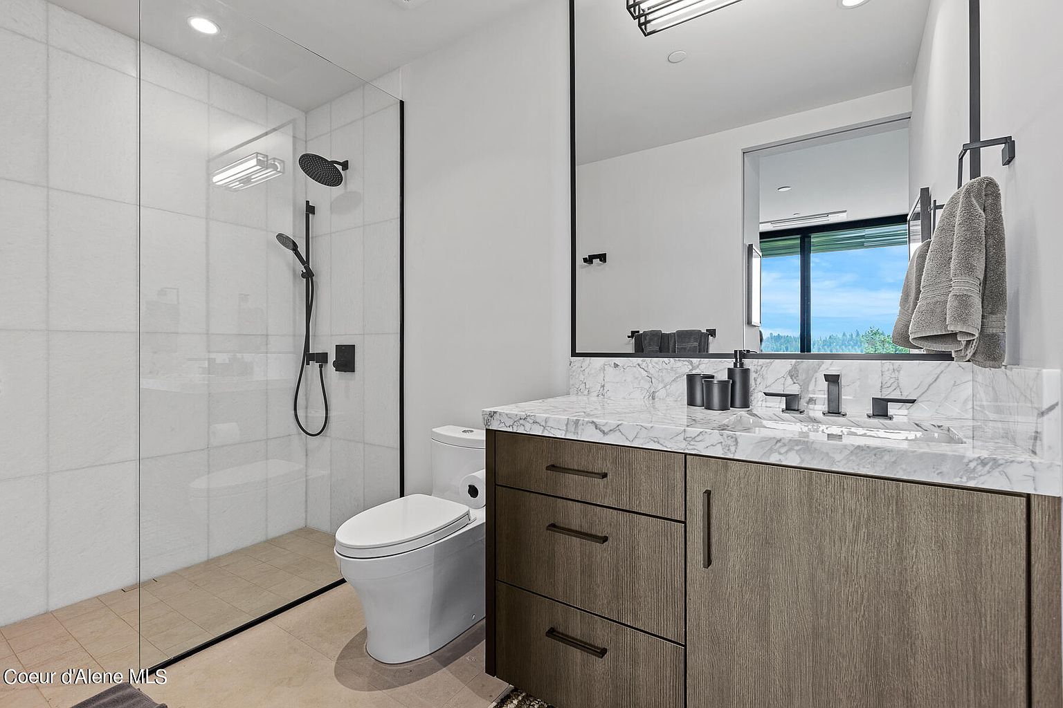This is a modern bathroom featuring a glass-enclosed shower with a black shower head and fixtures, a white toilet, and a vanity with a marble countertop and dark wood cabinets. A large mirror hangs above the vanity, reflecting natural light from a nearby window. The overall design is sleek and minimalist.