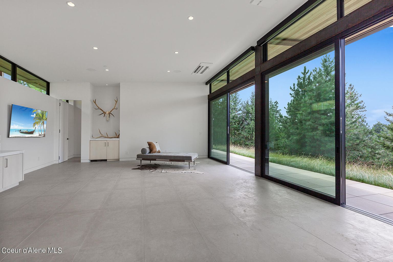 This is a spacious, modern living room with large sliding glass doors that offer a view of the surrounding greenery. The room features neutral-toned tile flooring, white walls, and minimalist furniture, including a daybed and a built-in cabinet. The overall impression is one of clean lines, natural light, and seamless integration with the outdoors.
