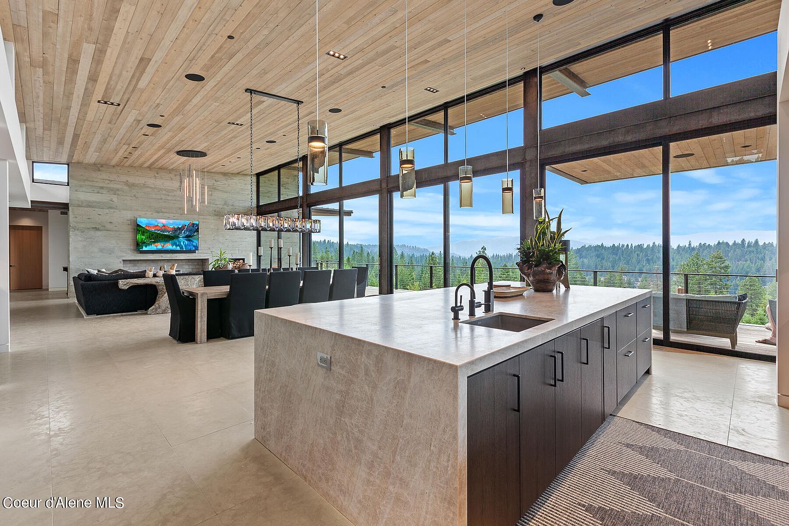 This interior shot showcases a modern, open-concept kitchen and dining area with floor-to-ceiling windows offering stunning views of the surrounding landscape. The kitchen features a large island with a light-colored countertop and dark cabinetry, while the dining area includes a long table with black chairs and a unique chandelier. The space is well-lit and exudes a sense of luxury and connection to nature.
