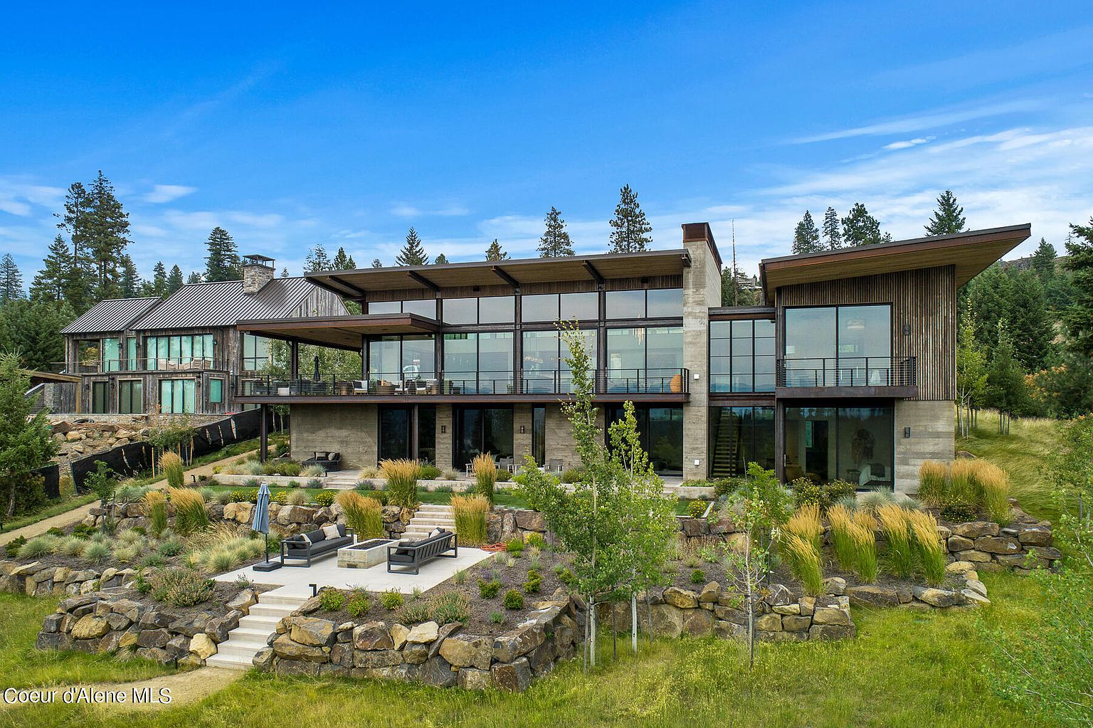 This image showcases the rear exterior of a modern luxury home, featuring a multi-level design with expansive glass windows and a flat roof. The architecture incorporates natural materials like wood and stone, blending seamlessly with the surrounding landscape. A patio area with outdoor seating and a fire pit is visible, enhancing the property's appeal for outdoor living and entertaining.
