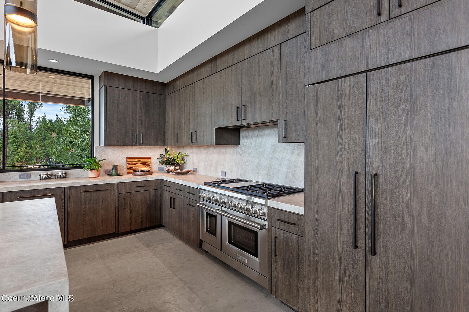 This is a modern kitchen featuring dark wood cabinetry and stainless steel appliances. The countertops are light-colored stone, and a large window provides natural light and a view of the outdoors. The kitchen has a sleek and sophisticated design.