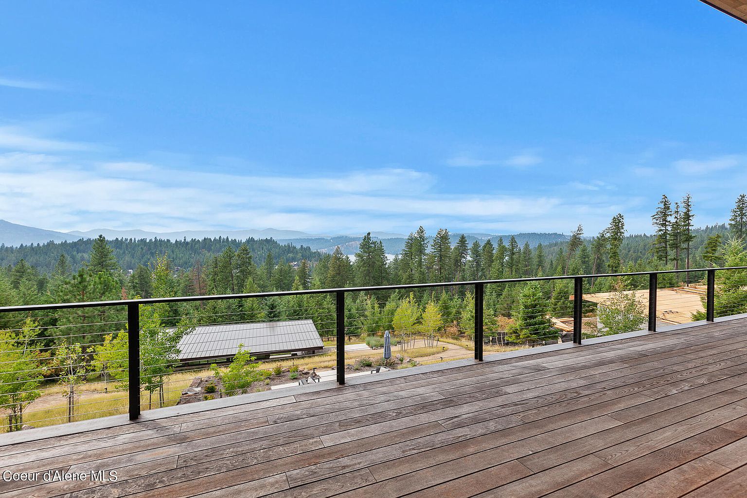 This image showcases a spacious wooden deck with a sleek, modern railing, offering panoramic views of a lush, forested landscape under a clear blue sky. The deck's warm wood tones contrast beautifully with the dark railing, creating an inviting outdoor living space perfect for relaxation and entertainment. A building with a metal roof is visible in the distance, adding to the property's appeal.