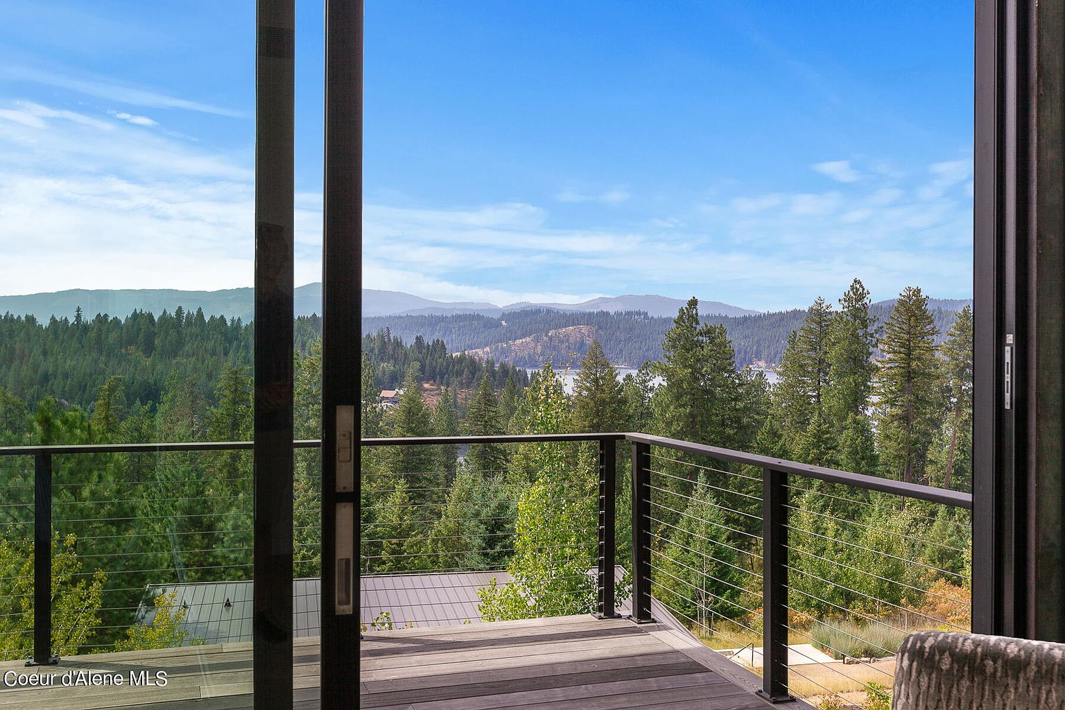 This image showcases a spacious balcony with a modern design, featuring a sleek railing and wooden decking. The view from the balcony is breathtaking, overlooking a lush forest, a serene lake, and distant mountains under a clear blue sky. The scene evokes a sense of tranquility and connection with nature, highlighting the property's desirable outdoor living space.