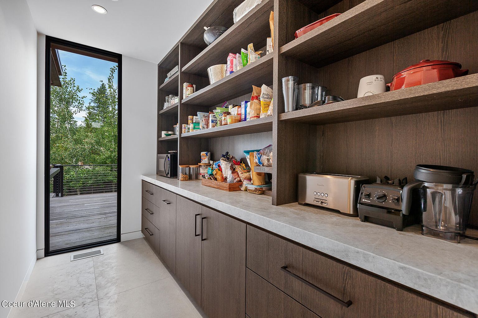 This image showcases a well-organized pantry with custom dark wood shelving and cabinetry, complemented by a light countertop. The pantry is stocked with various food items and appliances, including a toaster and blender. A large window provides natural light and a view of the outdoors, enhancing the space's appeal.